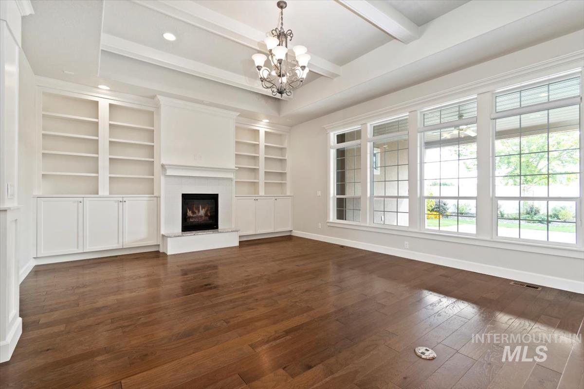 Unfurnished living room featuring beam ceiling, dark wood-type flooring, a warm lit fireplace, built in features, and a chandelier