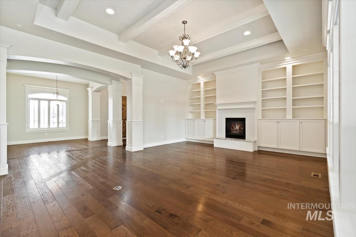 Unfurnished living room with beam ceiling, a chandelier, arched walkways, a warm lit fireplace, and ornate columns