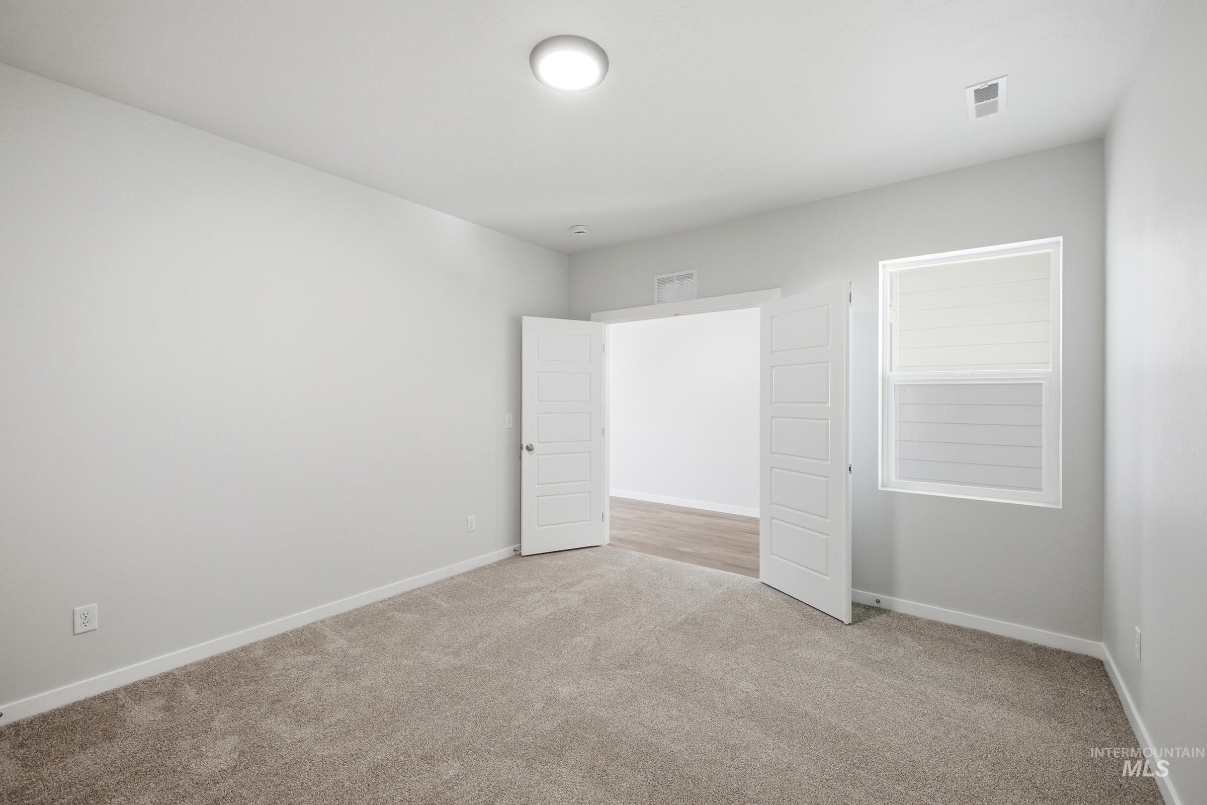 Spacious room featuring neutral carpeting, light gray wall tones, a modern ceiling light fixture, a window with horizontal blinds, and double paneled doors opening to an additional area with wood-finish flooring
