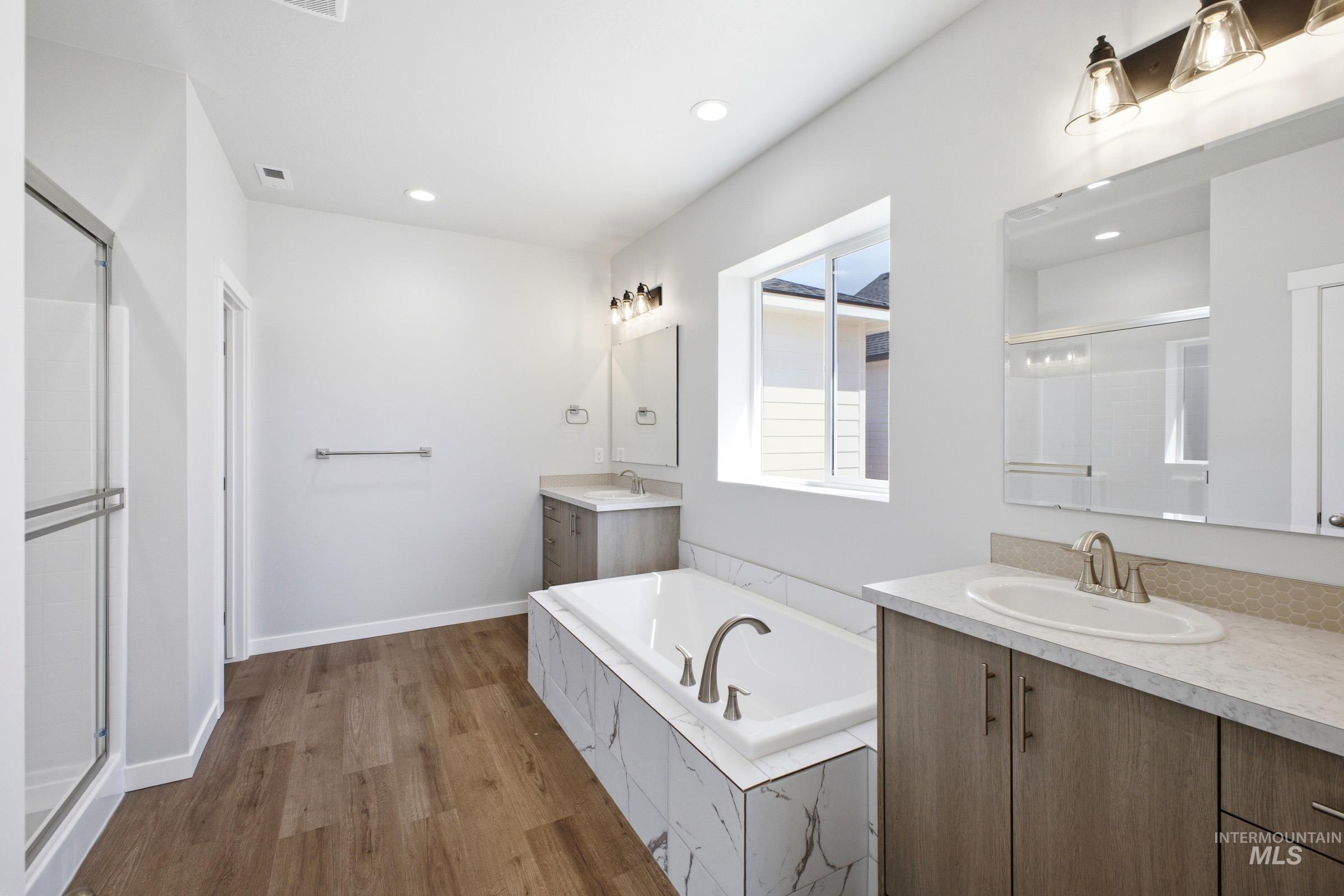 Contemporary bathroom featuring a large soaking tub with marble-look tile surround, dual vanities with integrated sinks, a separate glass-enclosed shower, and wood-finish flooring