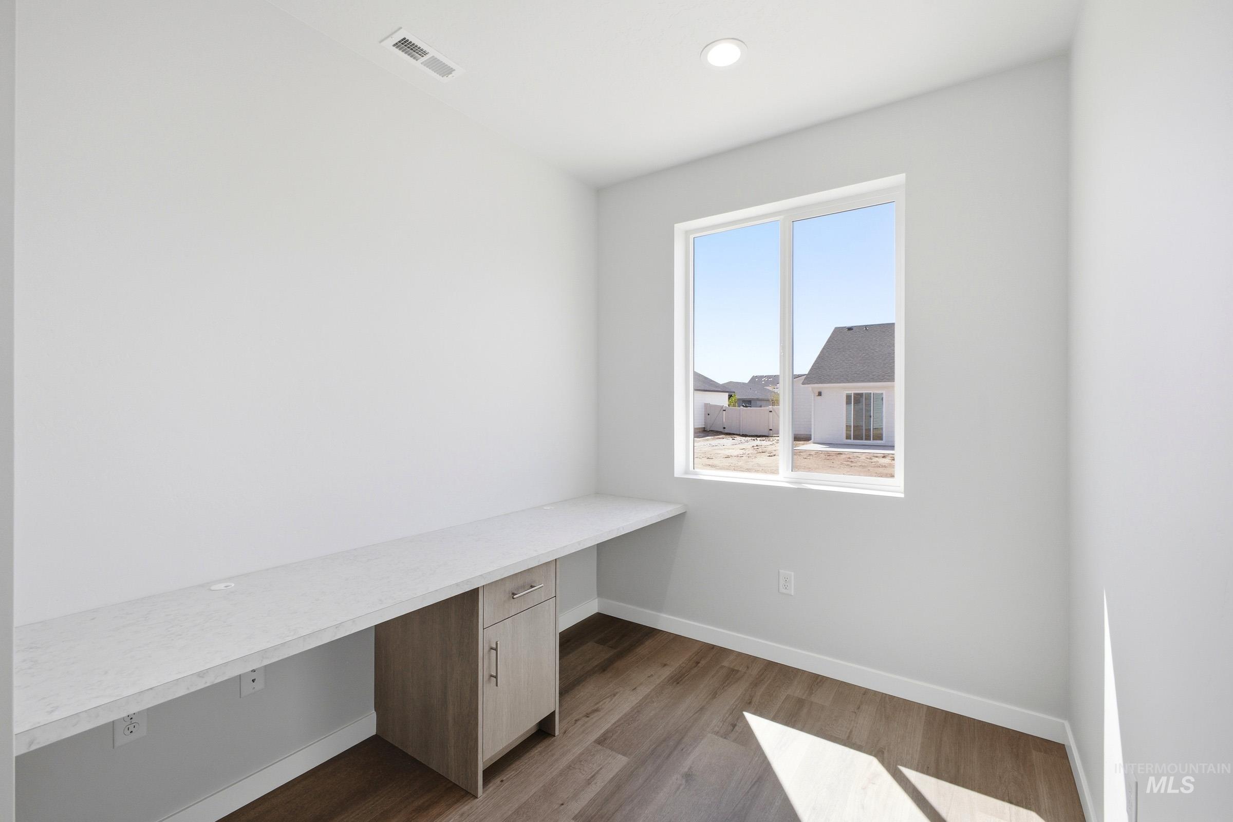 Bright room featuring a large window, built-in desk with a light-toned countertop, wood-finish flooring, recessed lighting, and a light wood-finish cabinet
