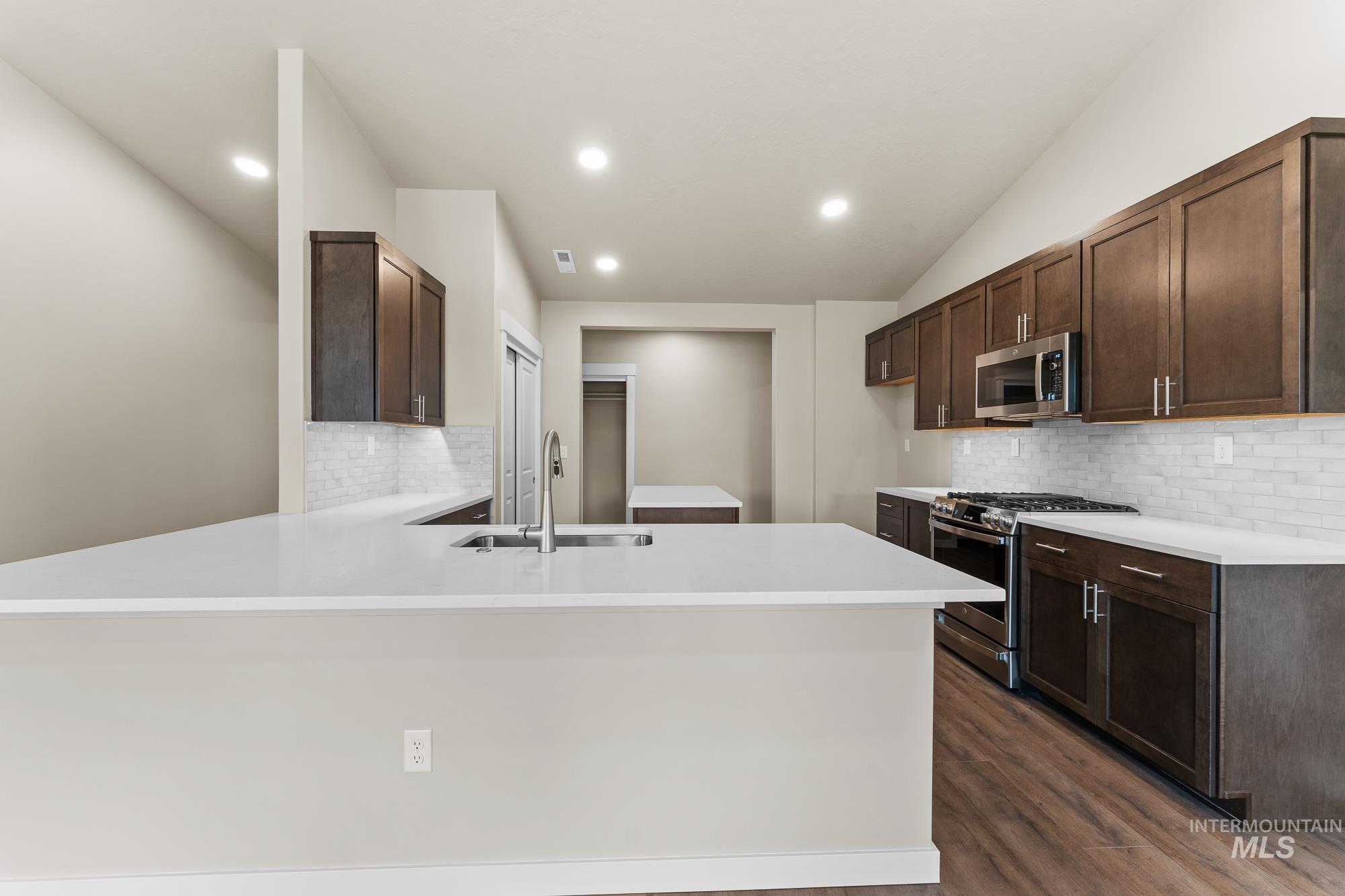 Spacious kitchen featuring an extended white countertop island, dark wood-finish cabinetry, stainless steel appliances, white subway tile backsplash, and wood-finish flooring