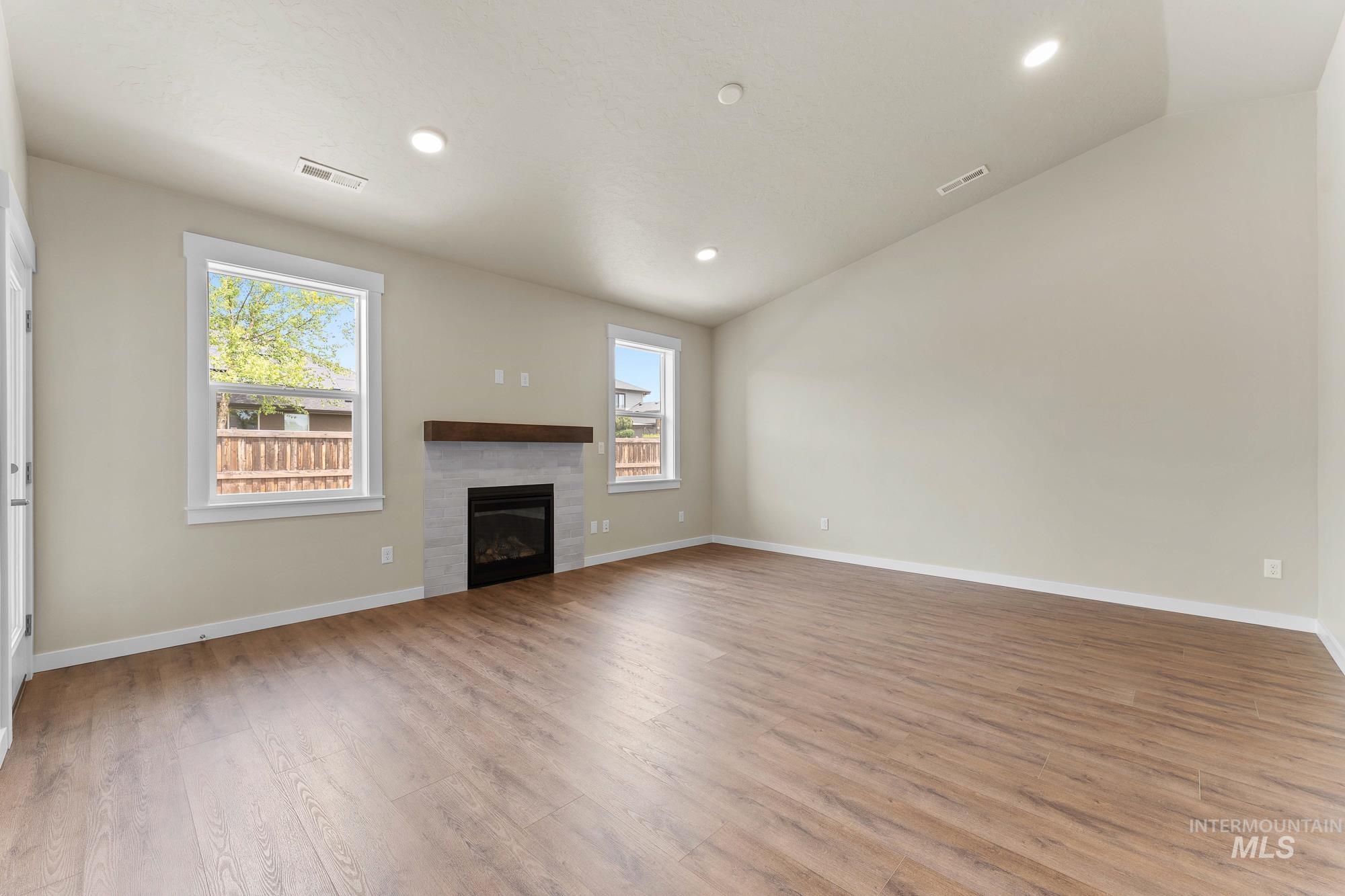 Spacious room featuring wood-finish flooring, a contemporary fireplace with a stone surround and wood mantel, multiple windows, and a vaulted ceiling with recessed lighting