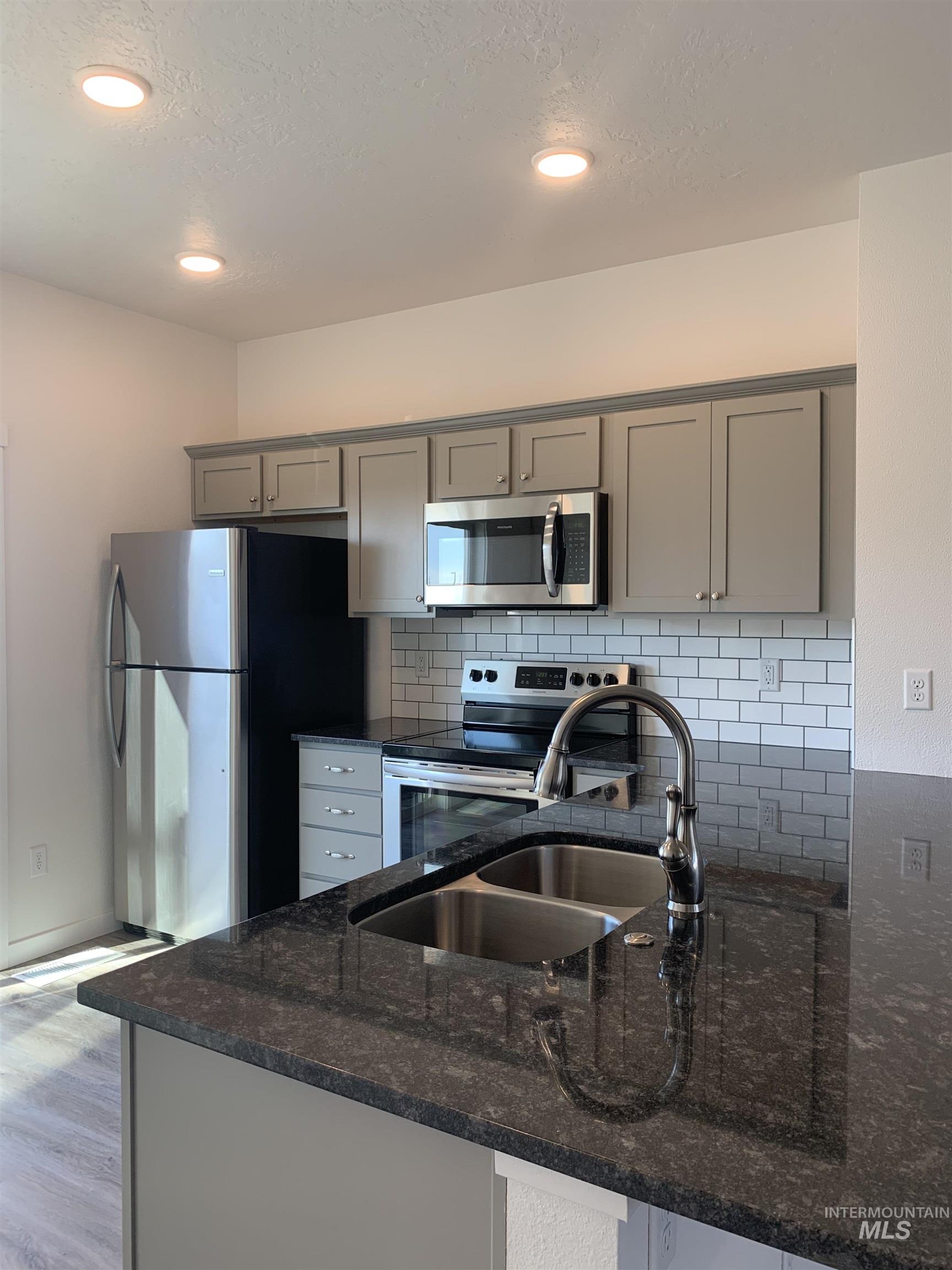 Kitchen featuring gray cabinetry, stainless steel appliances, dark stone counters, decorative backsplash, and light wood finished floors
