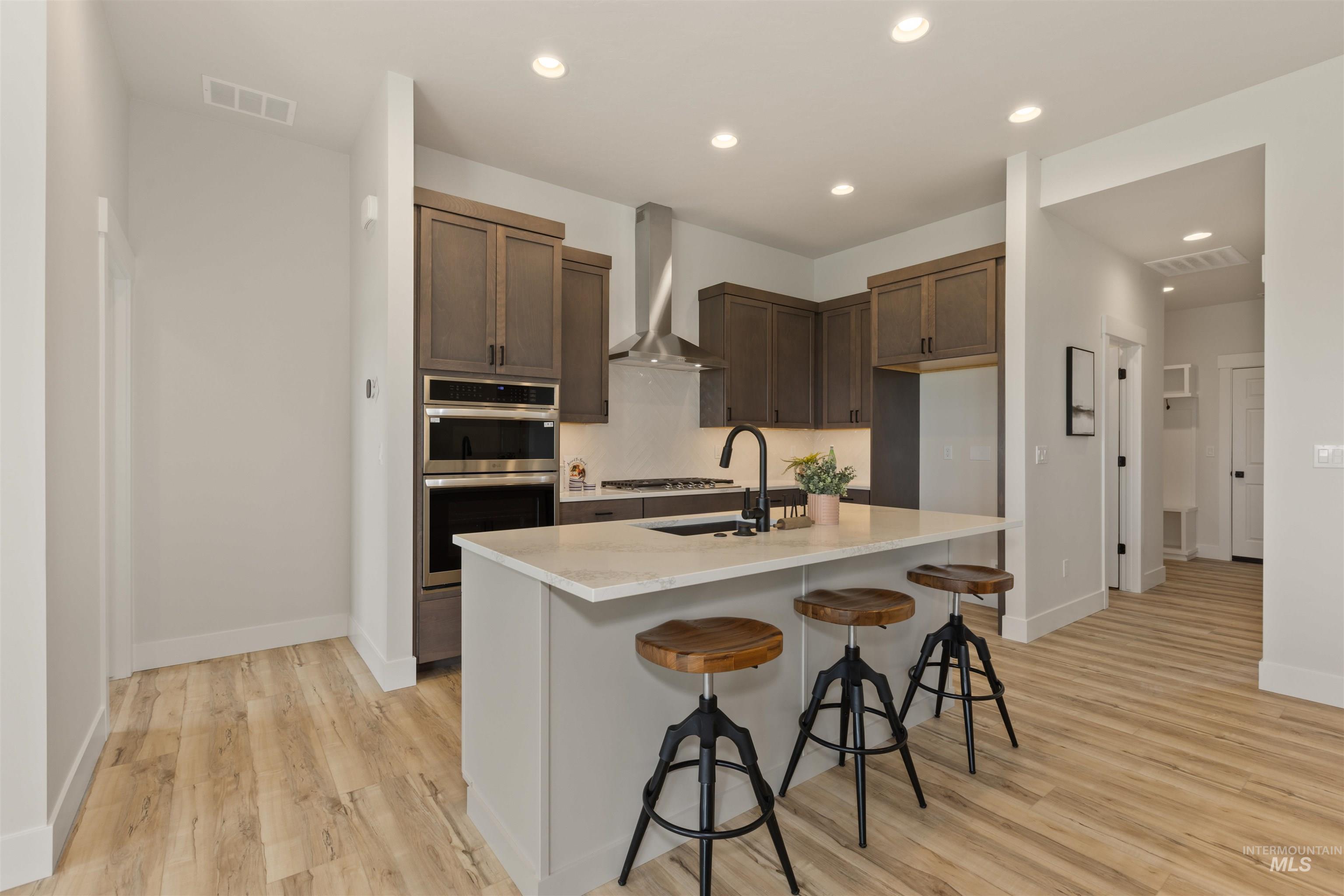 Kitchen featuring recessed lighting, wall chimney range hood, light wood-style floors, a breakfast bar area, and a kitchen island with sink