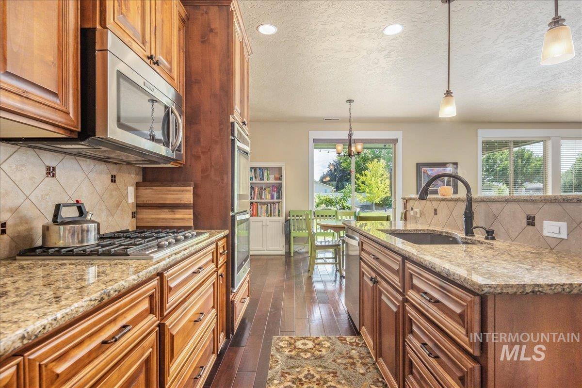 Kitchen with backsplash, brown cabinetry, appliances with stainless steel finishes, light stone counters, and a textured ceiling