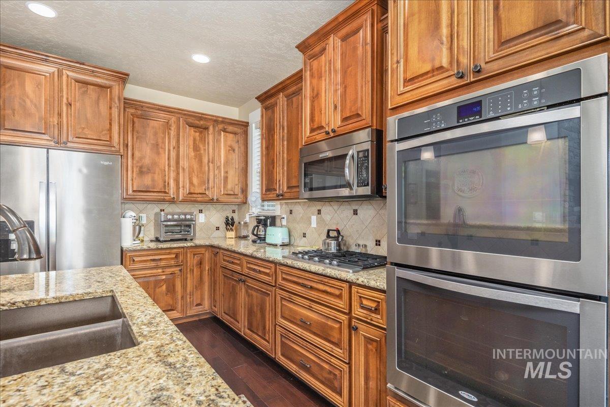 Kitchen featuring stainless steel appliances, decorative backsplash, light stone counters, brown cabinets, and a textured ceiling