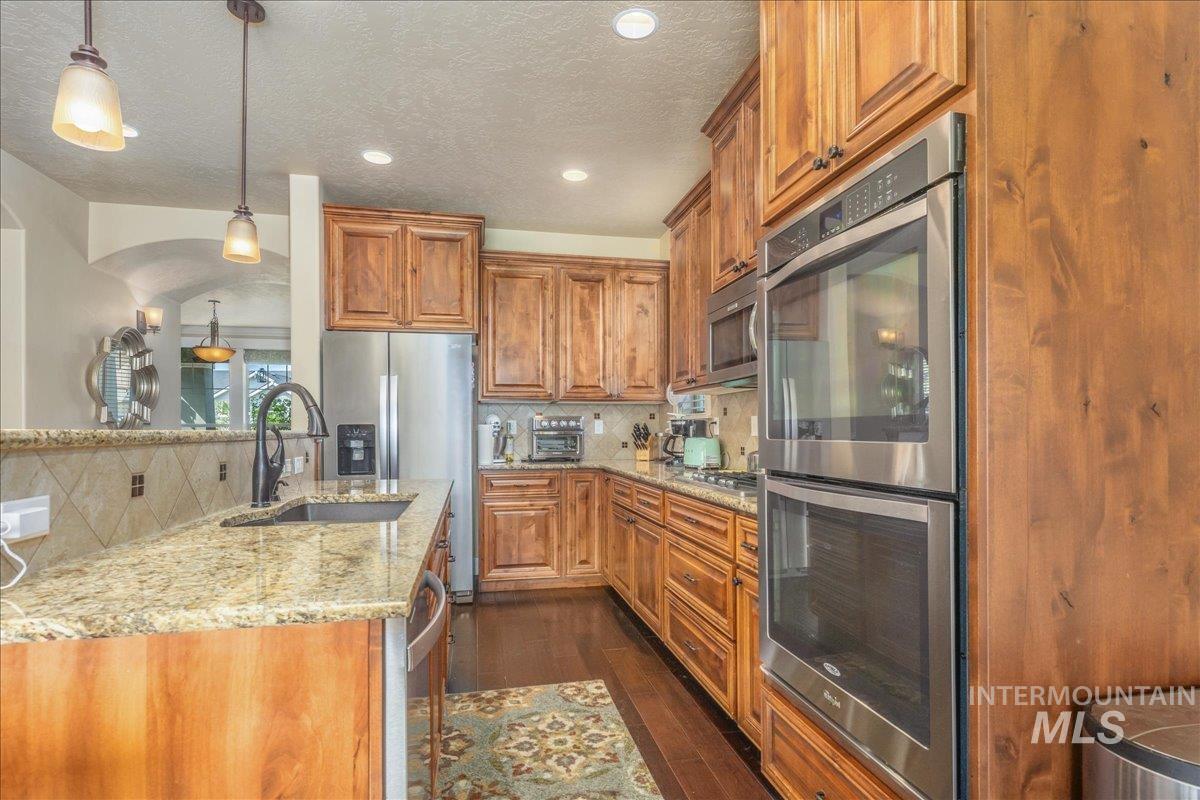 Kitchen with appliances with stainless steel finishes, decorative backsplash, brown cabinetry, dark wood-style flooring, and light stone countertops