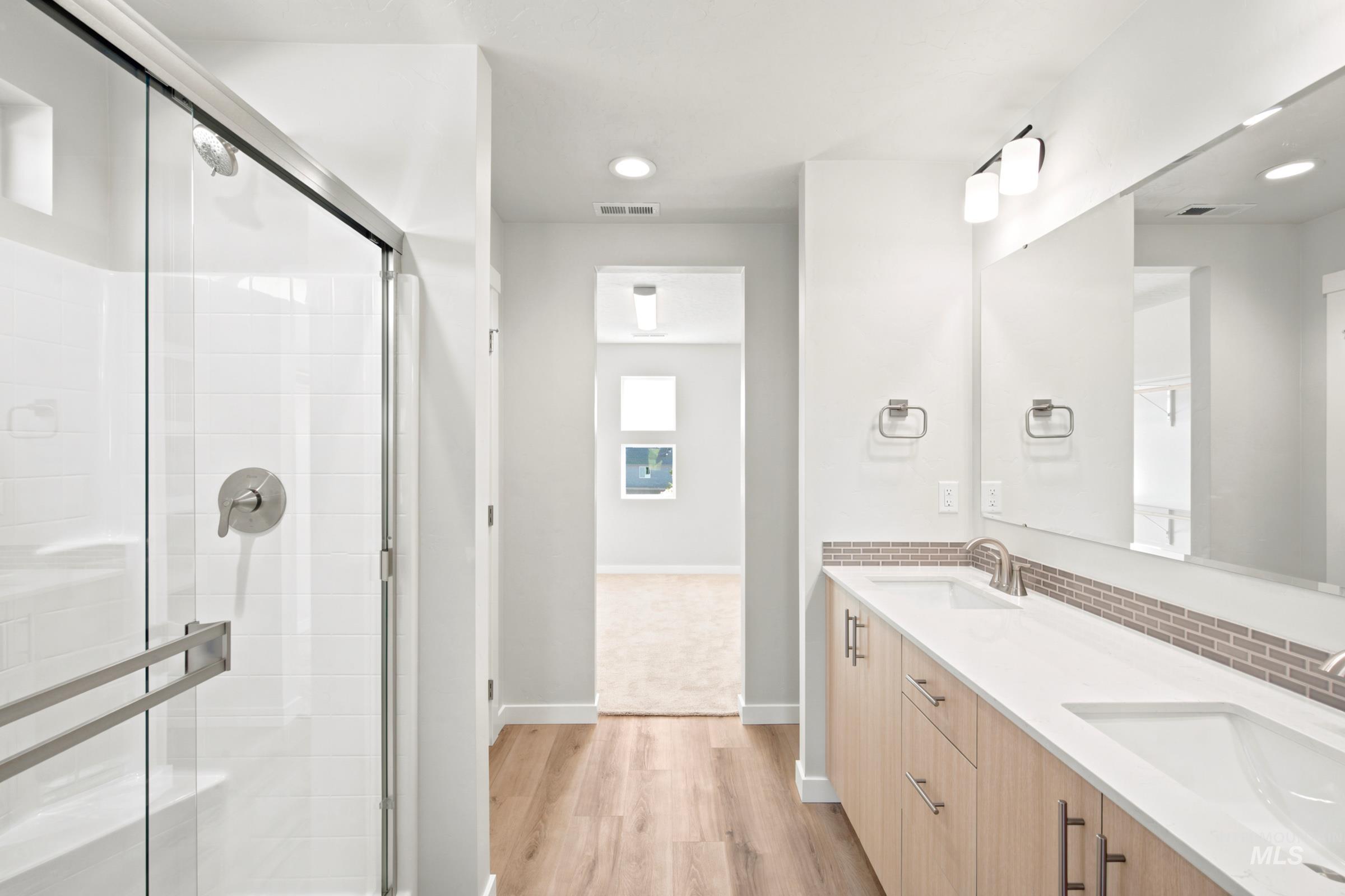 Modern bathroom featuring a glass-enclosed shower with white subway tile, a dual vanity with light wood-finish cabinetry, a white countertop, and a tiled backsplash