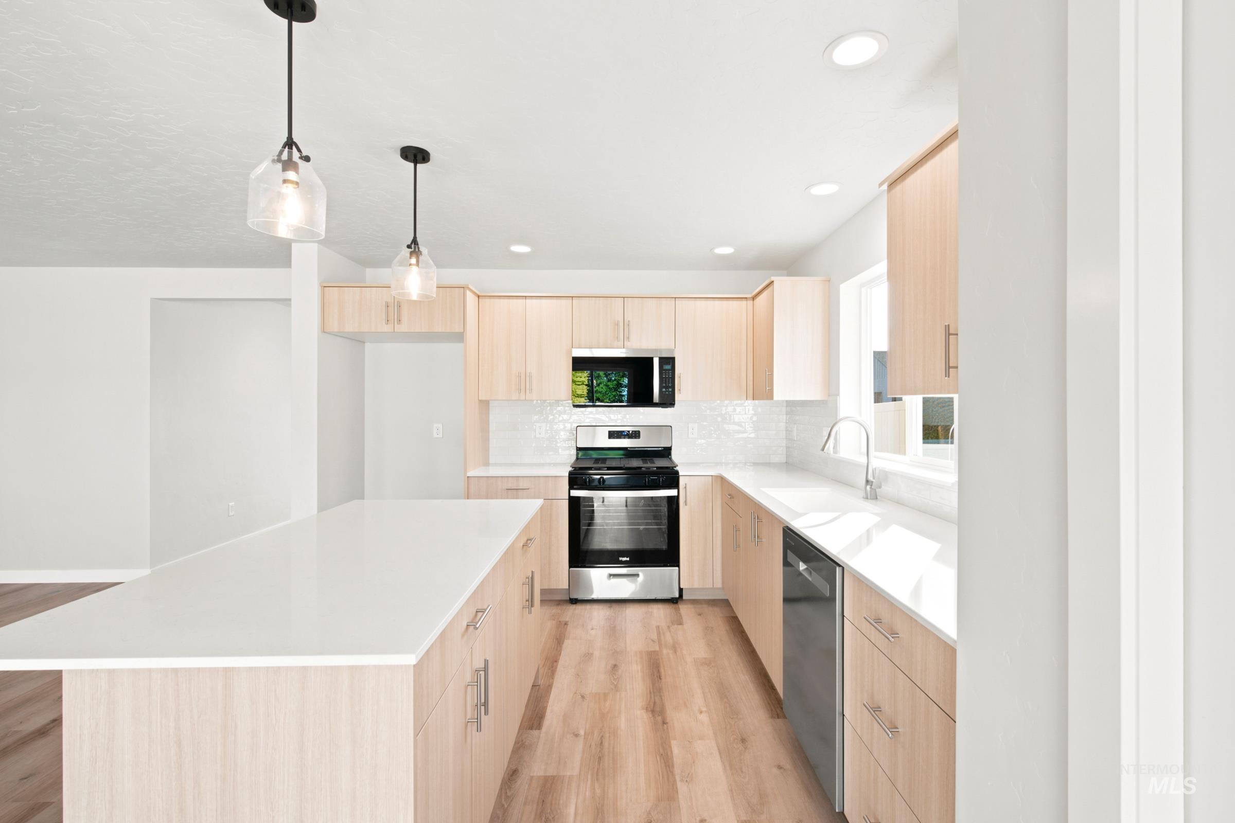 Spacious kitchen featuring a large island with pendant lighting, light wood-finish cabinetry, stainless steel appliances, white countertops, and wood-finish flooring