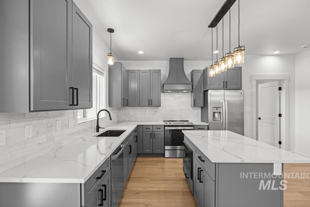 Kitchen with gray cabinetry, appliances with stainless steel finishes, tasteful backsplash, premium range hood, and light wood-style flooring