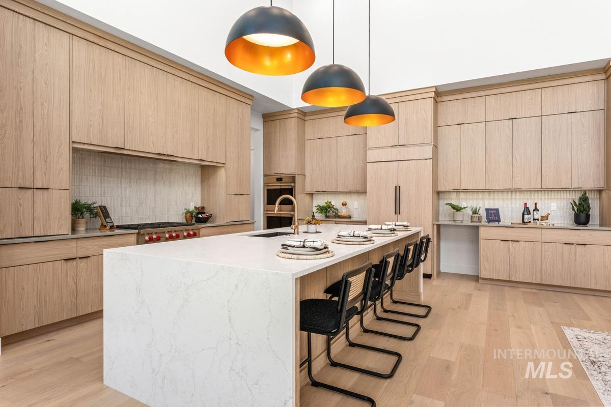 Spacious kitchen featuring an oversized island with countertop seating, light wood-finish cabinetry, a built-in range with a tile backsplash, and three contemporary pendant lights