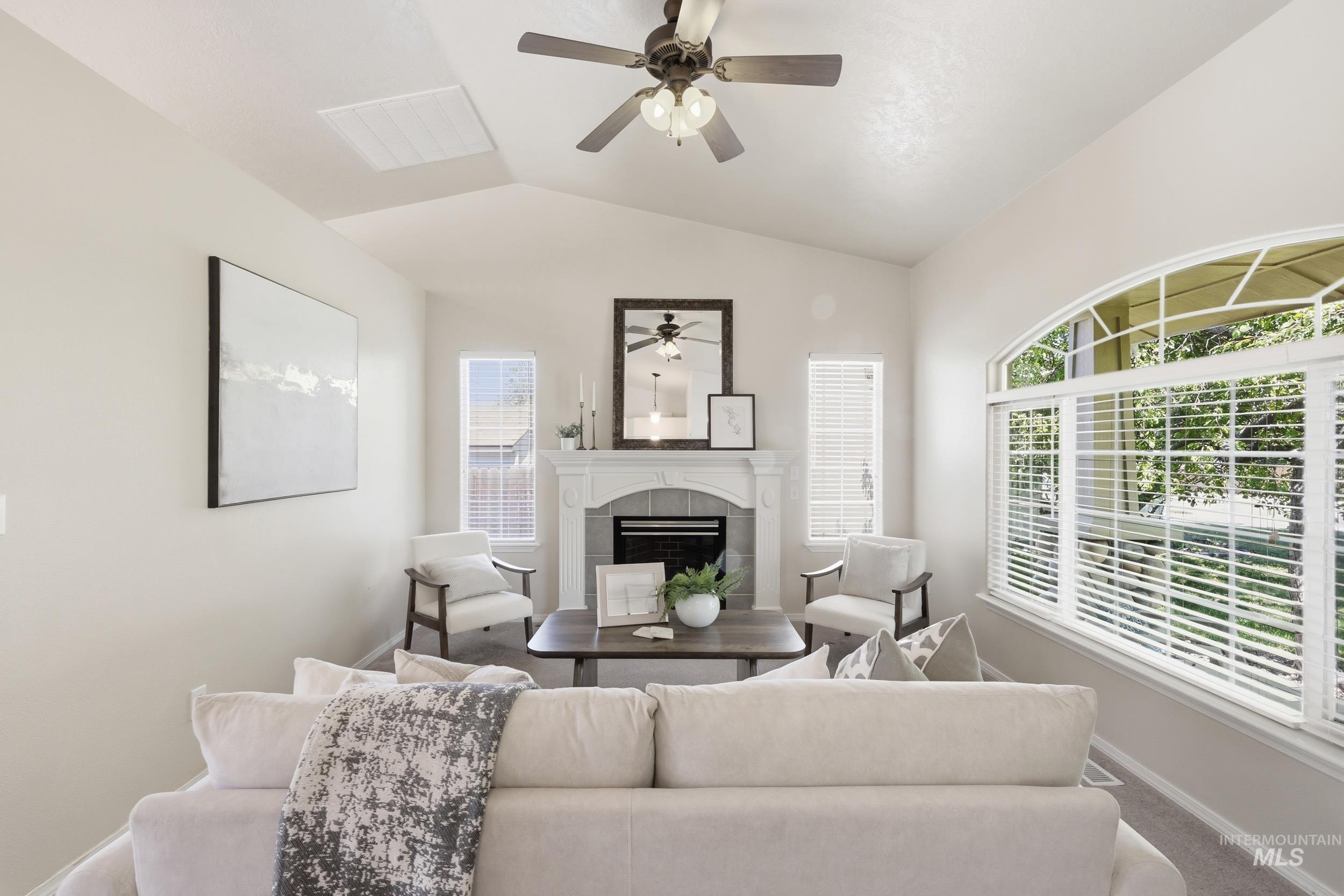 Living area featuring a vaulted ceiling with a ceiling fan, a fireplace with a white mantel, and a large arched window with horizontal blinds