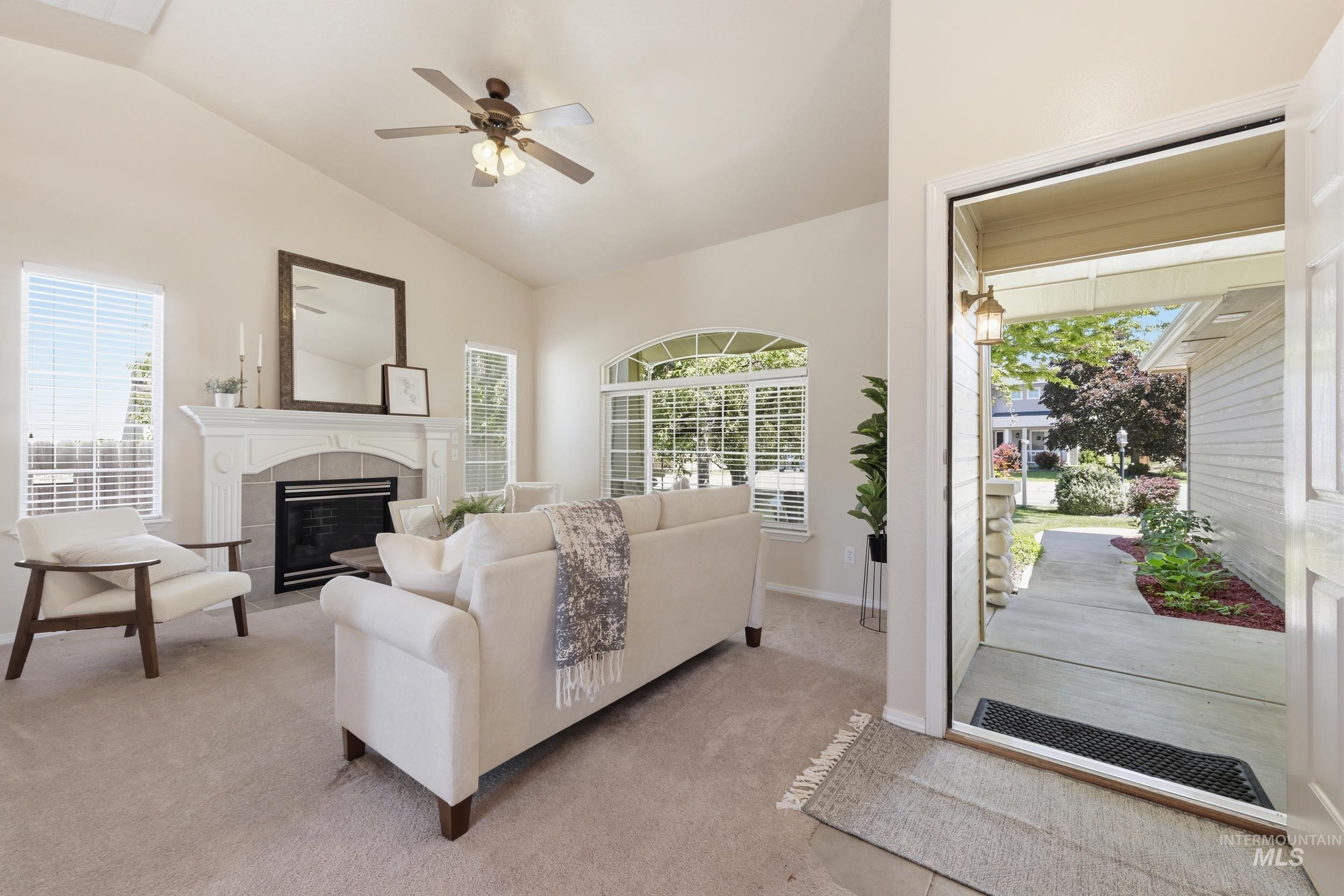 Vaulted ceiling living room featuring a decorative fireplace with a mantel, arched window, ceiling fan, and light-colored carpet flooring