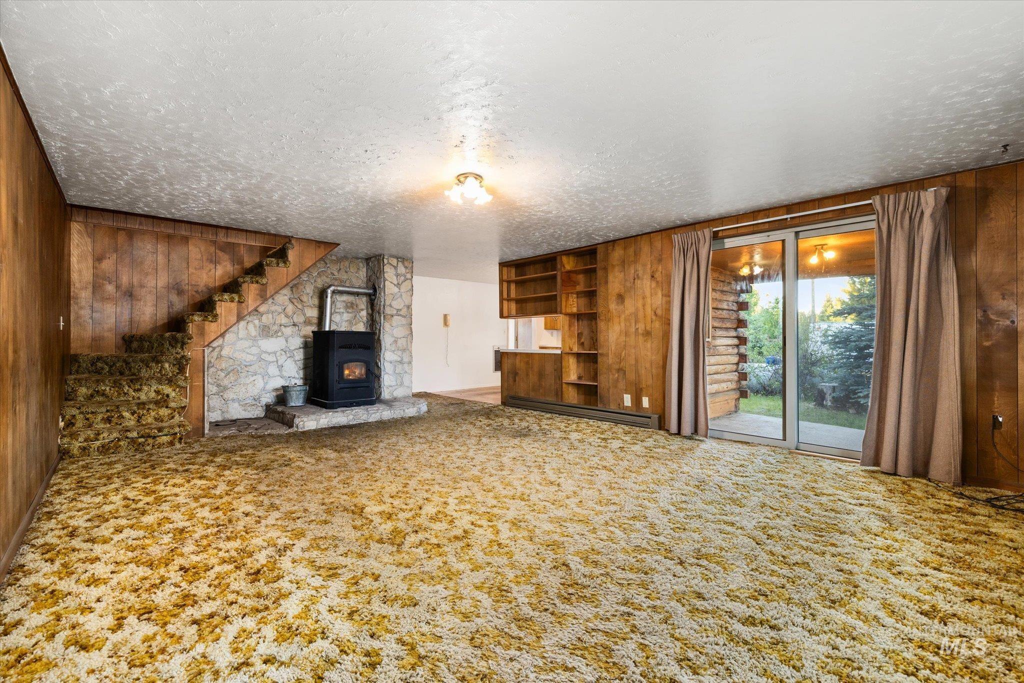 Spacious room featuring wood paneling, a stone-faced hearth with a wood-burning stove, built-in shelving, and a carpeted staircase