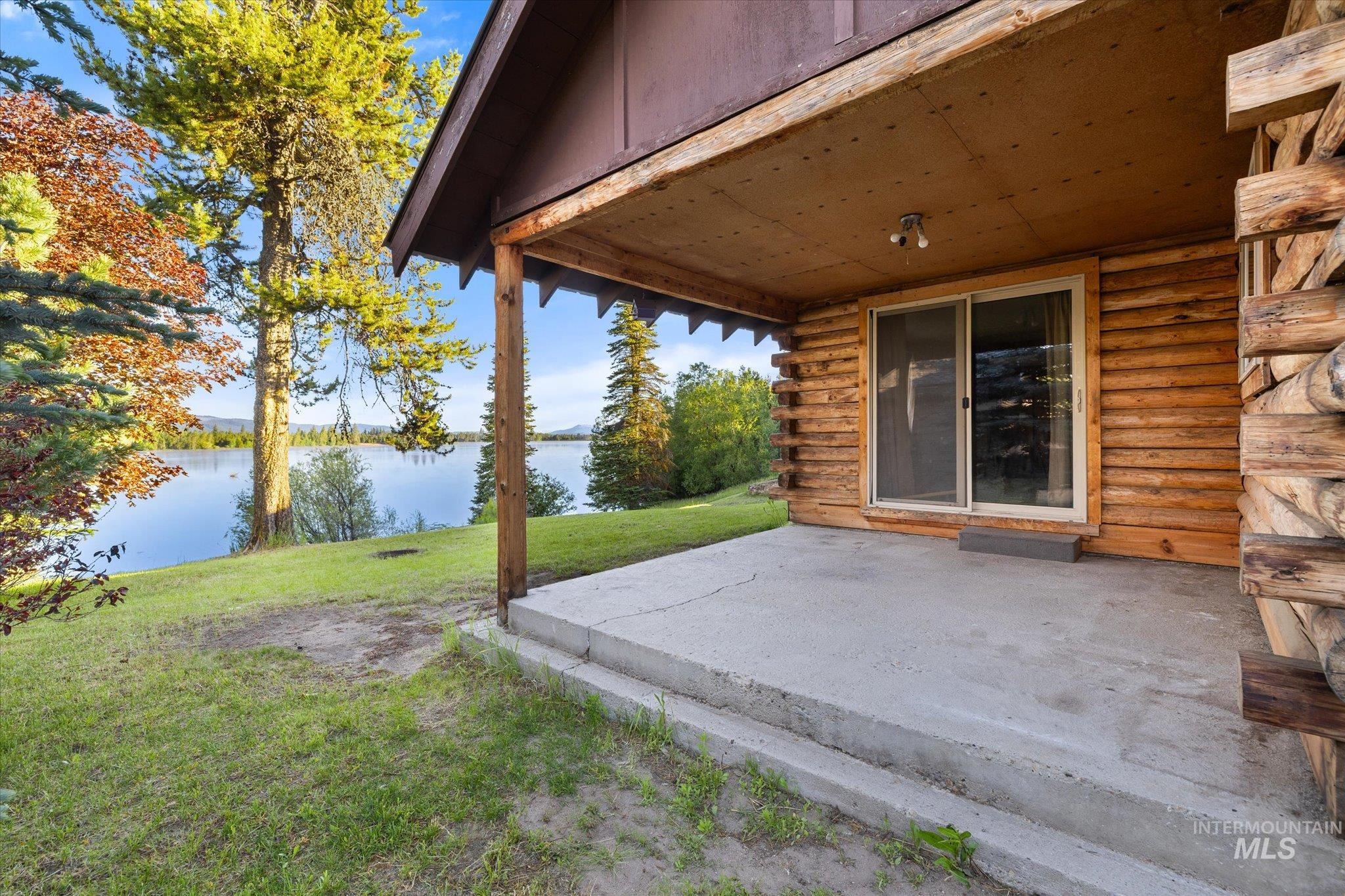 Covered concrete patio with log siding, a sliding glass door, and overhead lighting, featuring a waterfront setting with mature trees and a grassy lawn