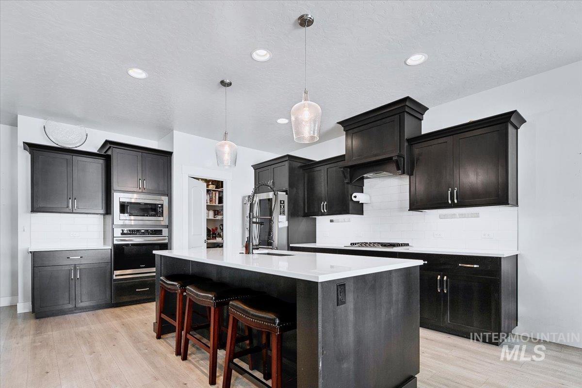 Modern kitchen featuring dark cabinetry, white subway tile backsplash, light wood-finish flooring, a central island with countertop seating, and recessed and pendant lighting