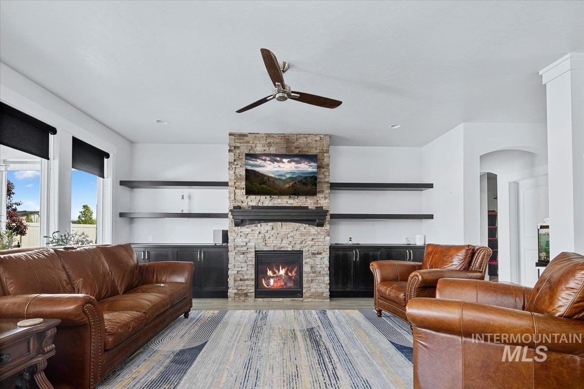 Spacious living area featuring a stone-clad fireplace with mantel, built-in cabinetry, floating shelving, and a ceiling fan