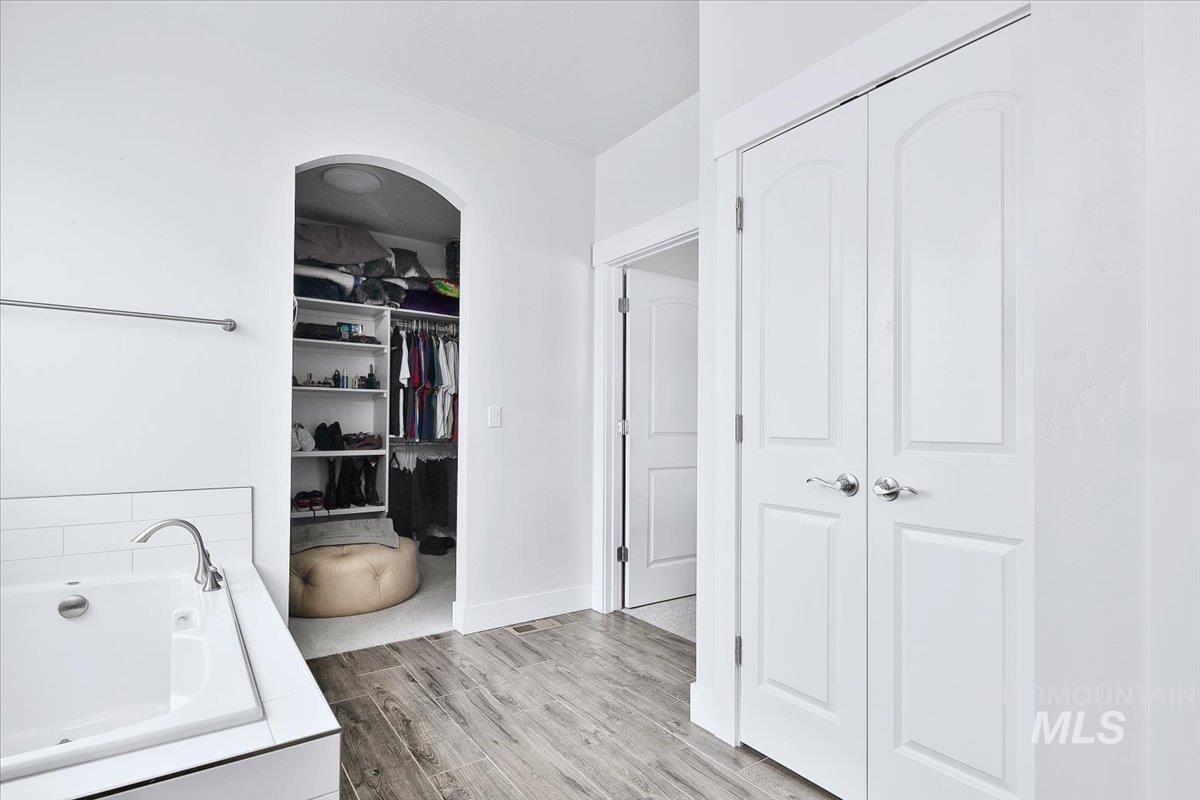 White soaking tub with subway tile surround, polished chrome faucet, wood-finish flooring, and an arched entry leading to a walk-in closet with built-in shelving