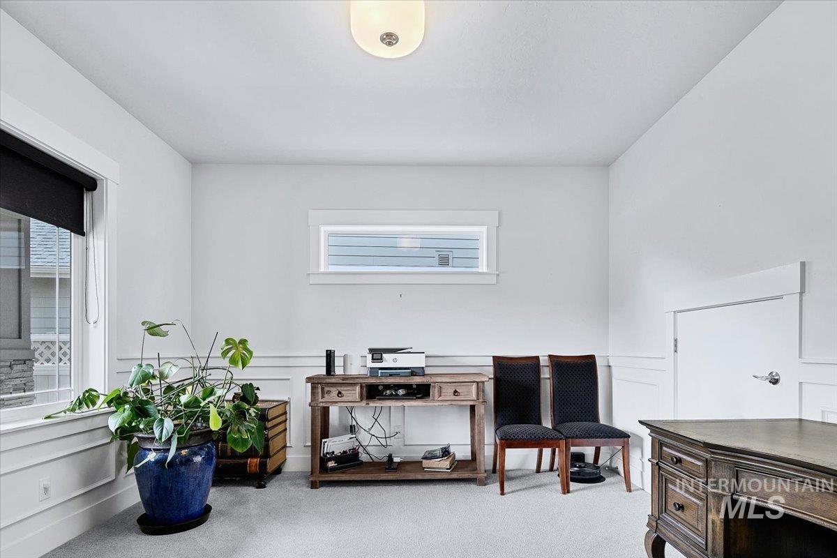 Bright room featuring a large window with roller blind, upper transom window, wainscoting details, light-toned carpet flooring, and a flush mount ceiling light