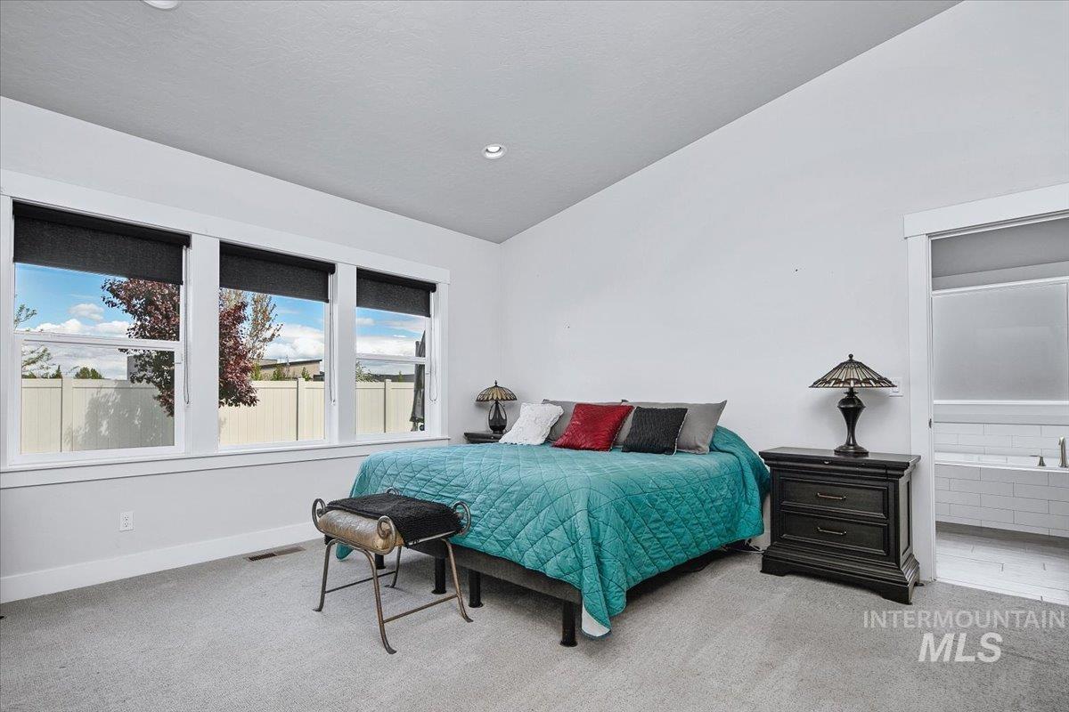 Spacious bedroom featuring multiple windows, vaulted ceilings, neutral-toned carpeting, and an adjoining bathroom with a built-in tub