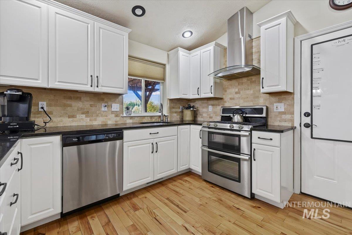 Kitchen with appliances with stainless steel finishes, white cabinets, wall chimney range hood, light wood finished floors, and decorative backsplash