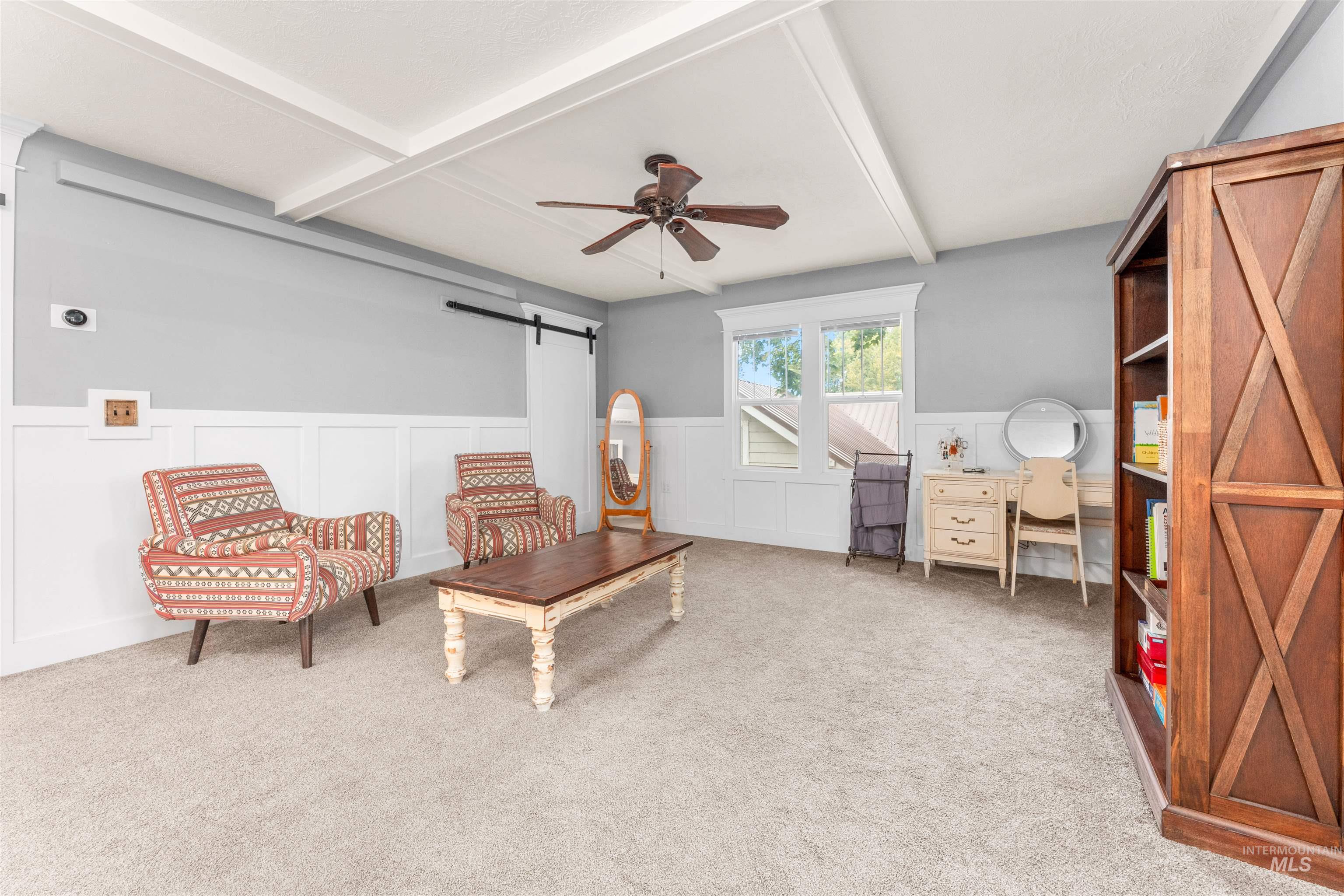 Living area featuring a decorative wall, a barn door, a wainscoted wall, a ceiling fan, and beamed ceiling