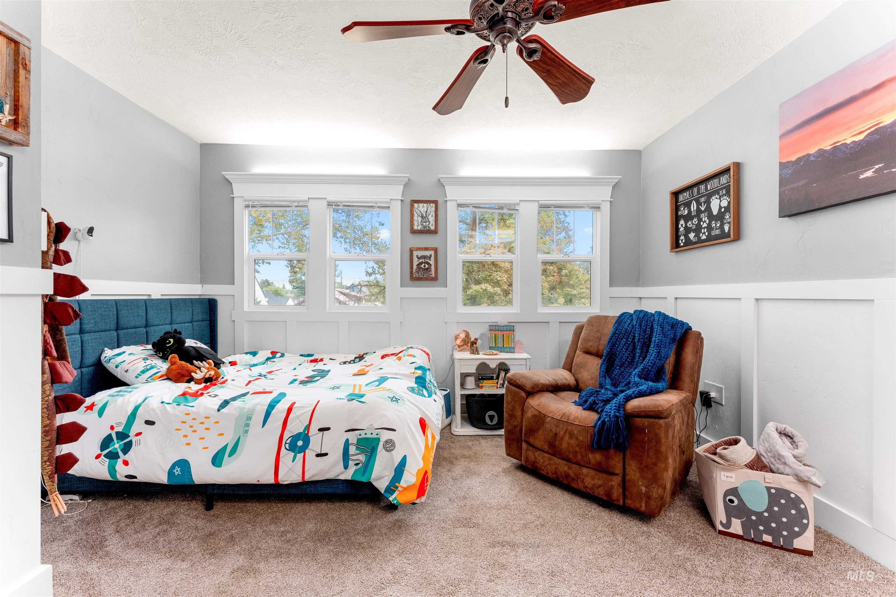Carpeted bedroom featuring a decorative wall, multiple windows, a wainscoted wall, a ceiling fan, and a textured ceiling