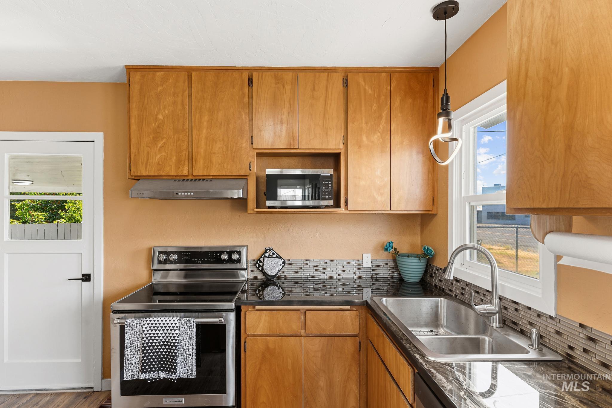 Kitchen featuring natural wood cabinetry, stainless steel appliances, dark stone countertops, a tile backsplash, and a double basin sink with a gooseneck faucet