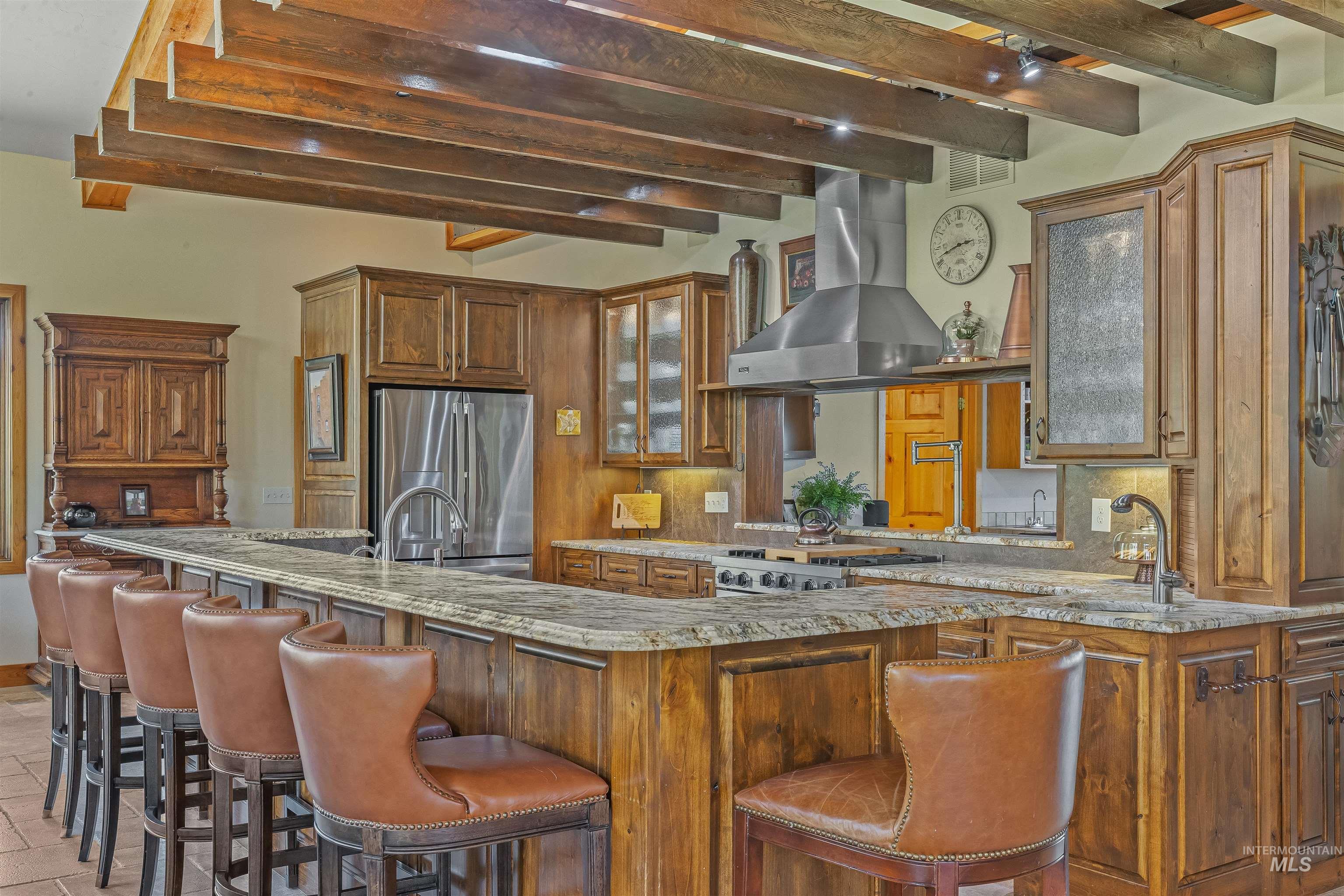 Kitchen with beamed ceiling, wall chimney range hood, backsplash, brown cabinets, and light stone counters
