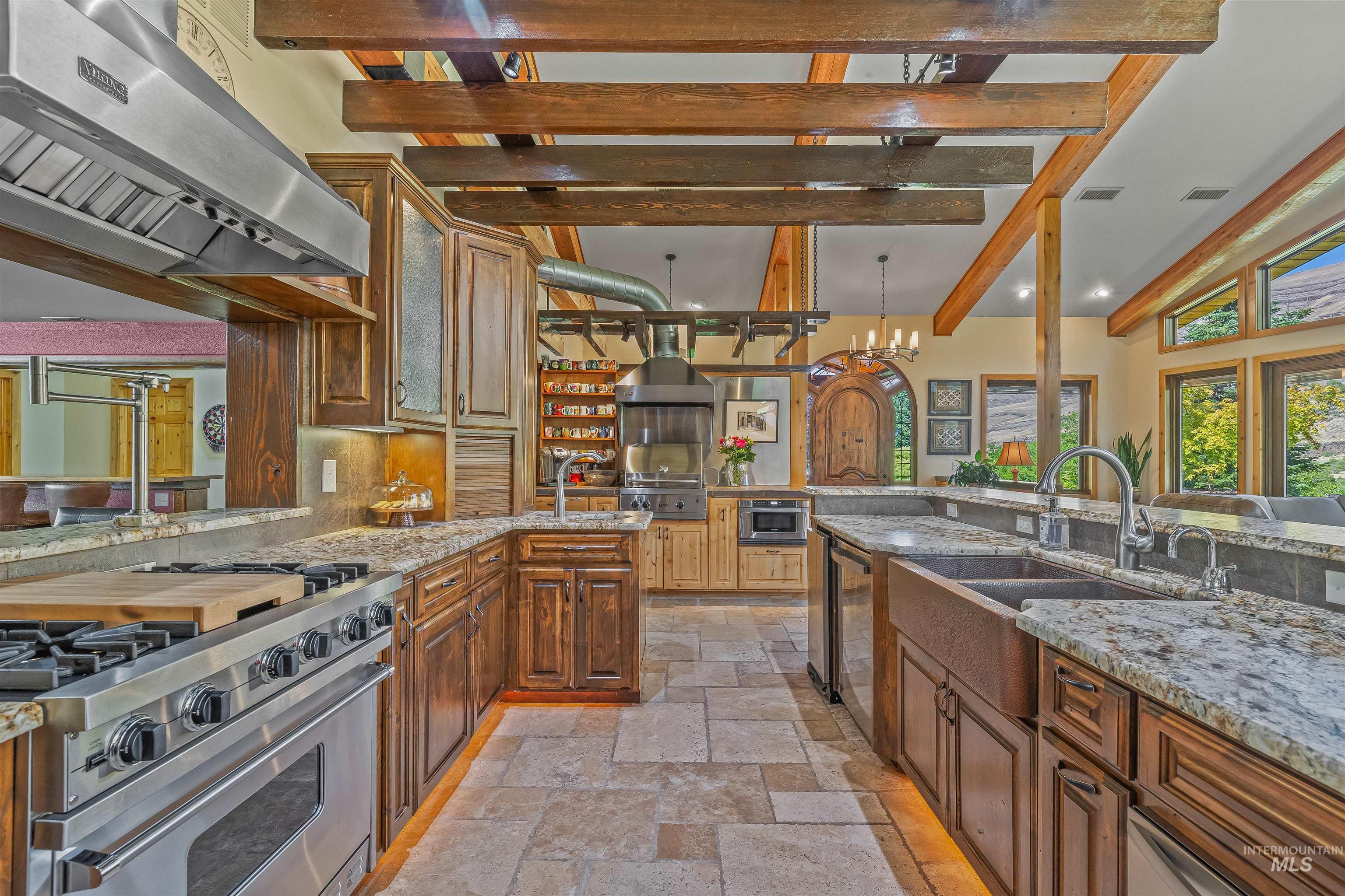 Kitchen with stone tile floors, appliances with stainless steel finishes, range hood, beamed ceiling, and decorative backsplash