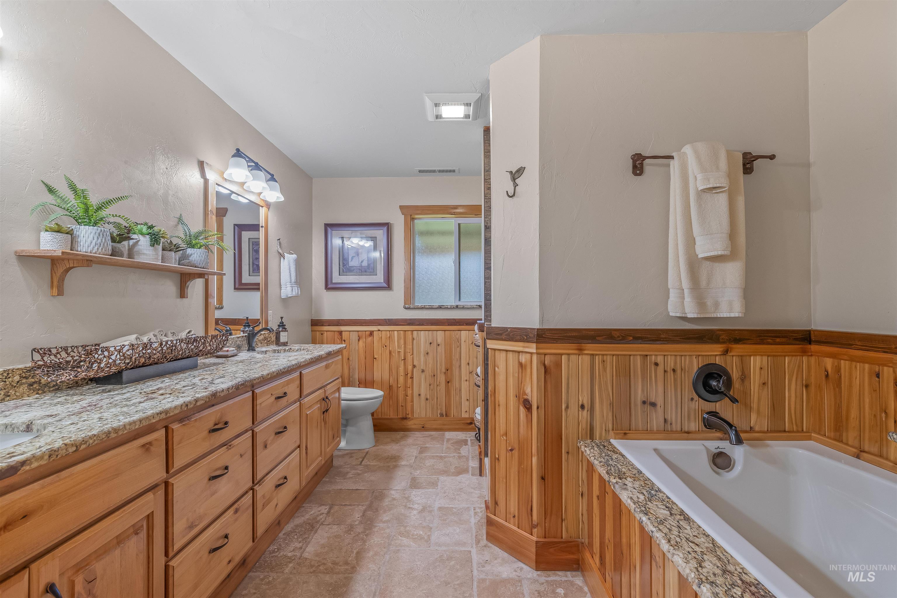 Full bathroom with a wainscoted wall, stone tile flooring, wood walls, double vanity, and a garden tub