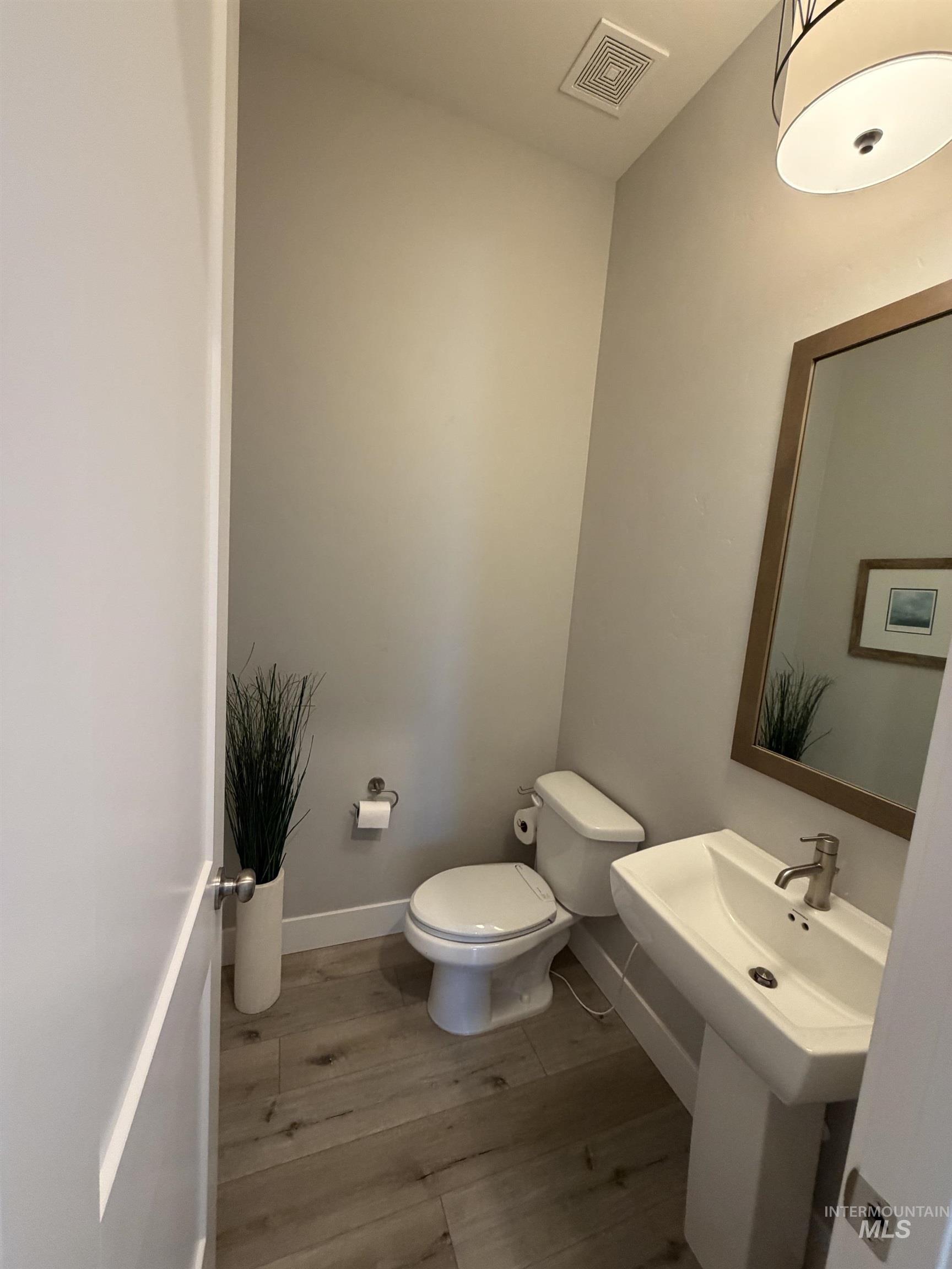 Powder room featuring wood-finish flooring, a contemporary pedestal sink, a toilet with a dual-flush mechanism, a wall-mounted mirror with a wood frame, and a modern drum pendant light fixture