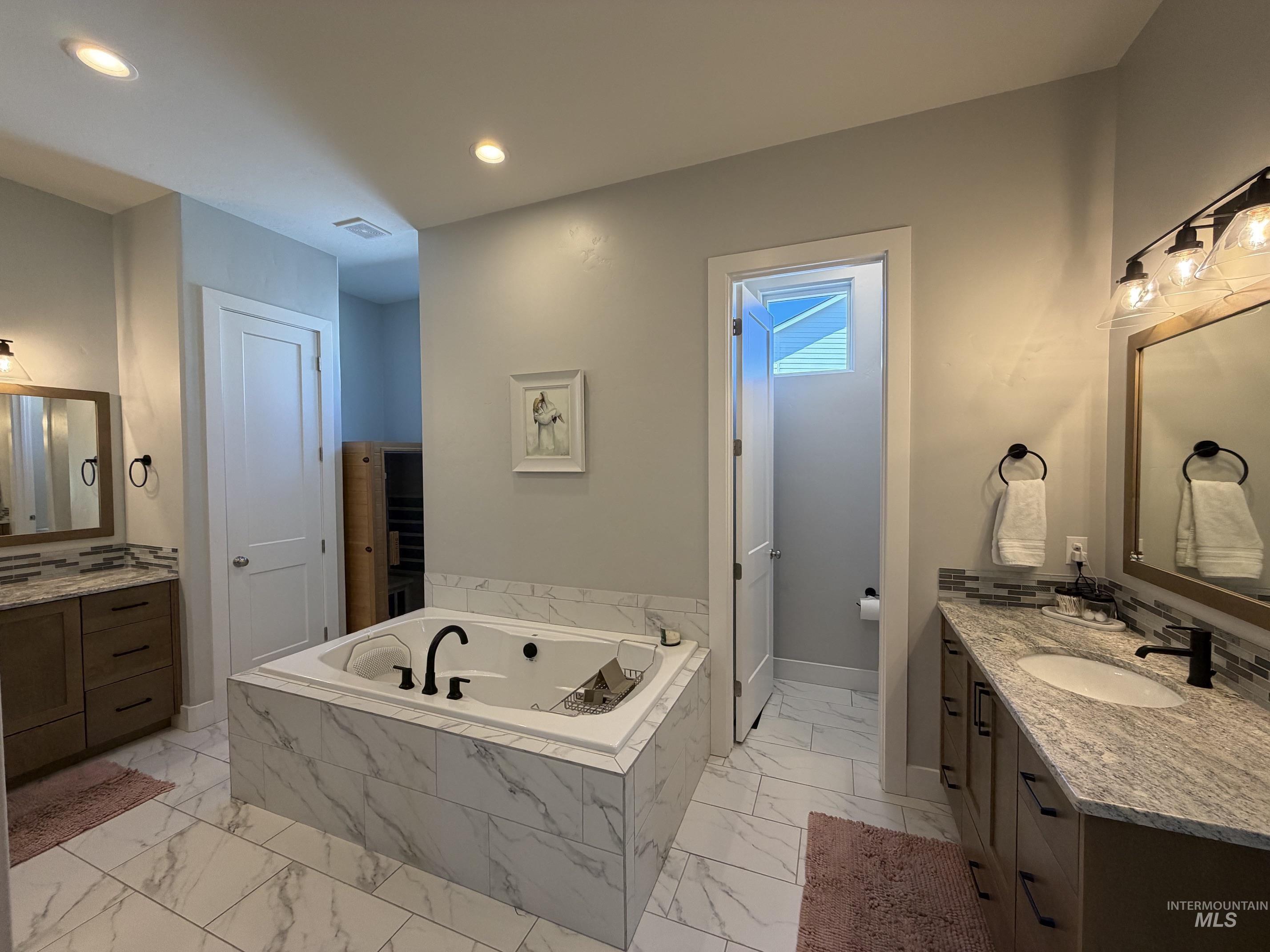 Spacious bathroom featuring a built-in soaking tub with marble-look tile surround, dual vanities with stone countertops, individual mirrors, and recessed lighting