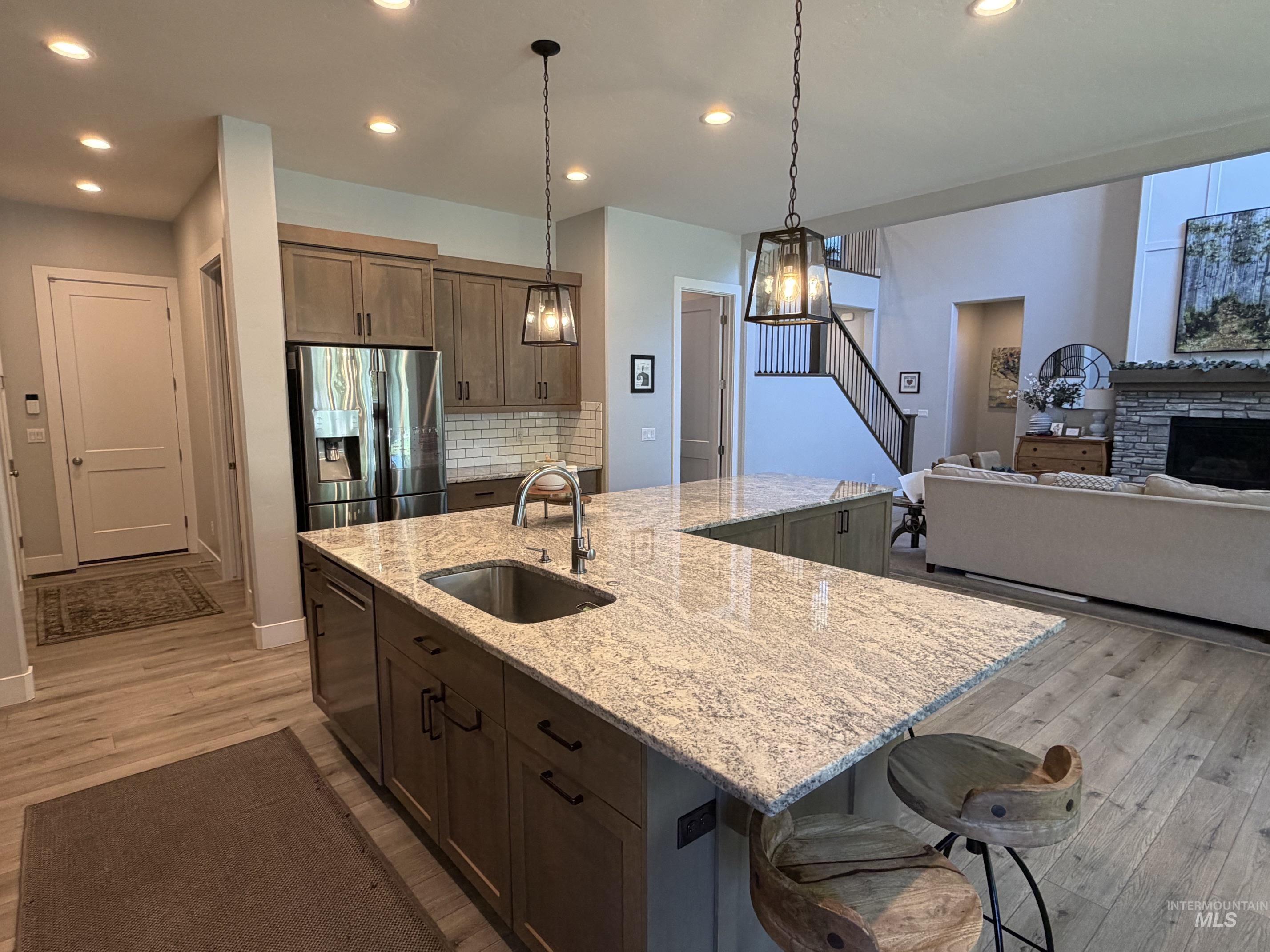 Open-concept living space featuring an L-shaped kitchen island with integrated sink, stainless steel appliances, wood-finish flooring, and a stone fireplace