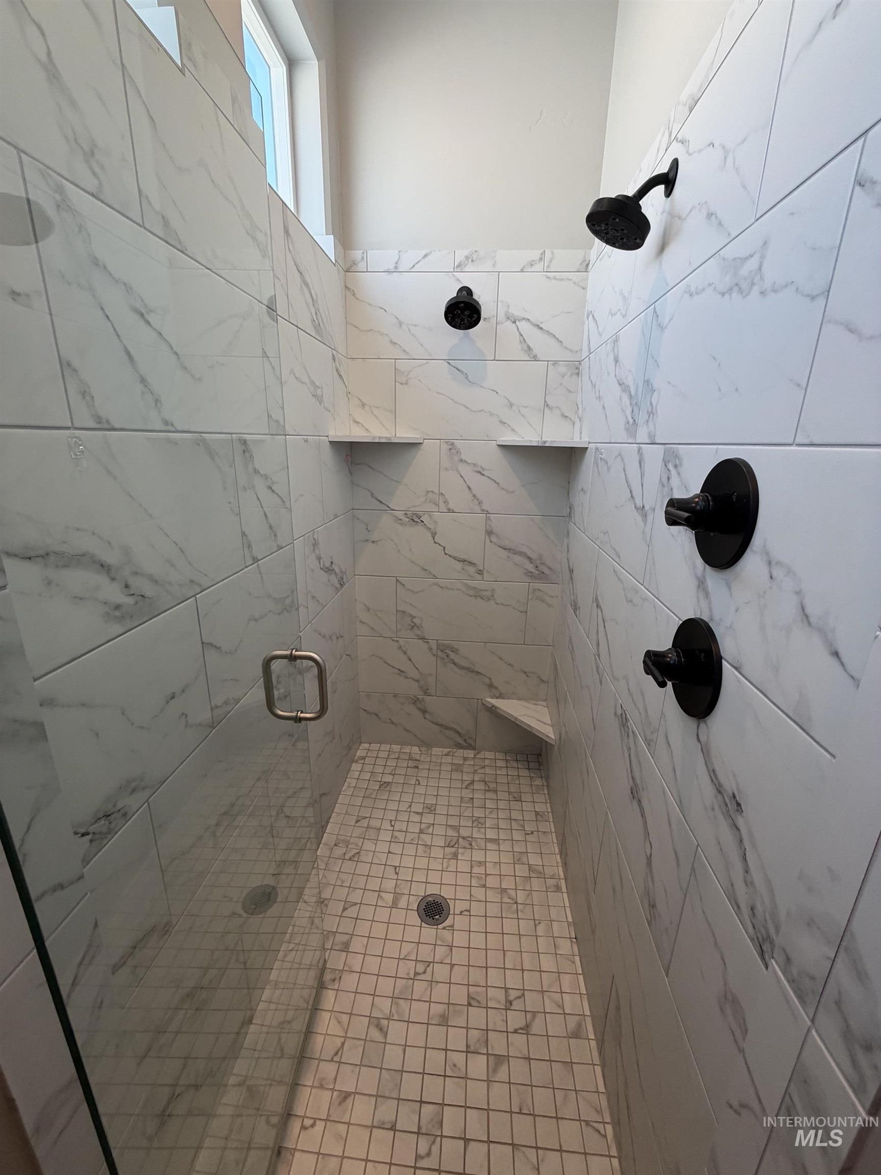 Spacious walk-in shower featuring large format white and gray marble-look wall tiles, mosaic floor tiles, and a clear glass enclosure with a gold-tone handle