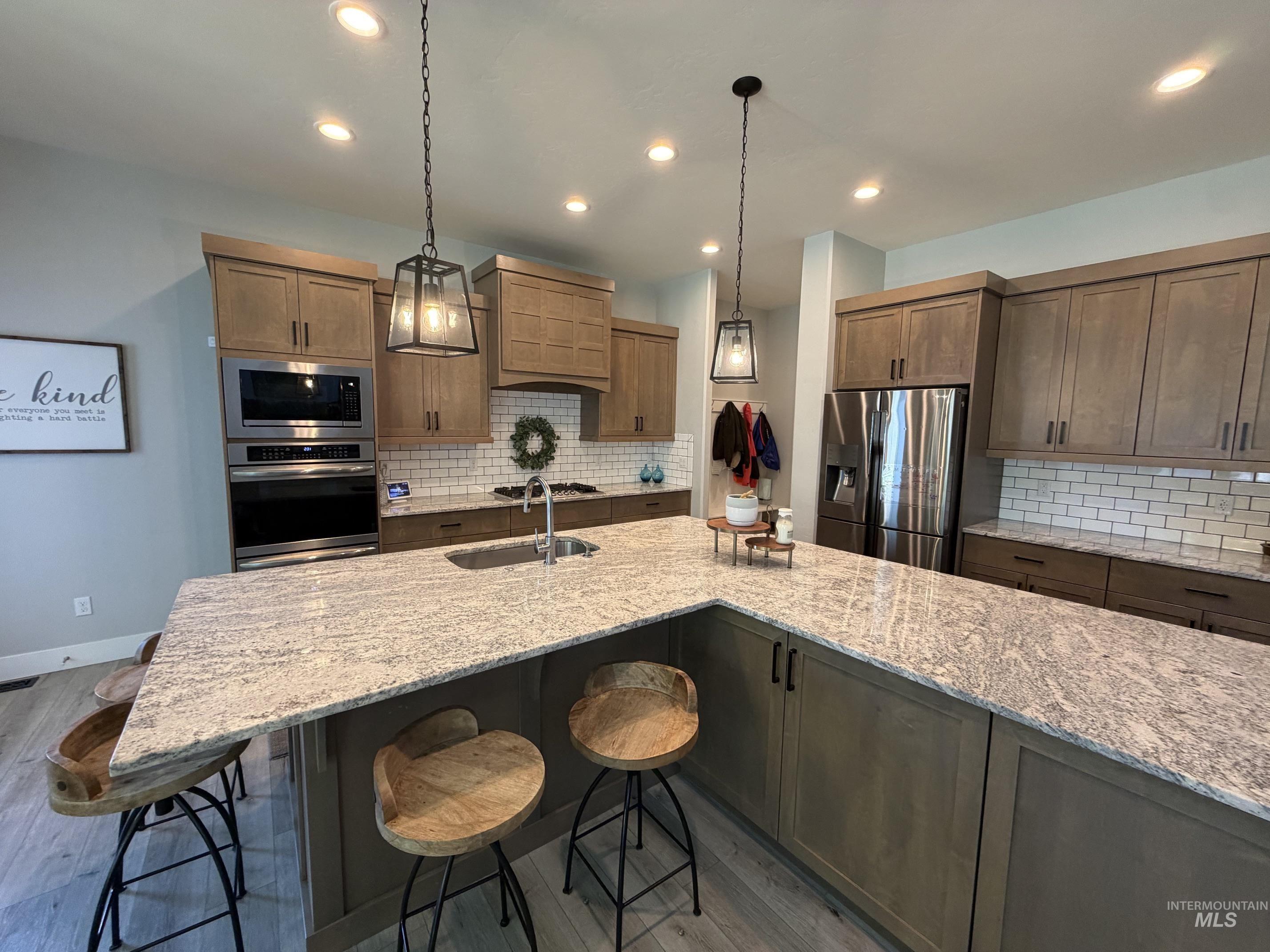 Expansive kitchen island featuring a light-toned stone countertop, coordinating subway tile backsplash, recessed lighting, stainless steel appliances, and wood-finish cabinetry