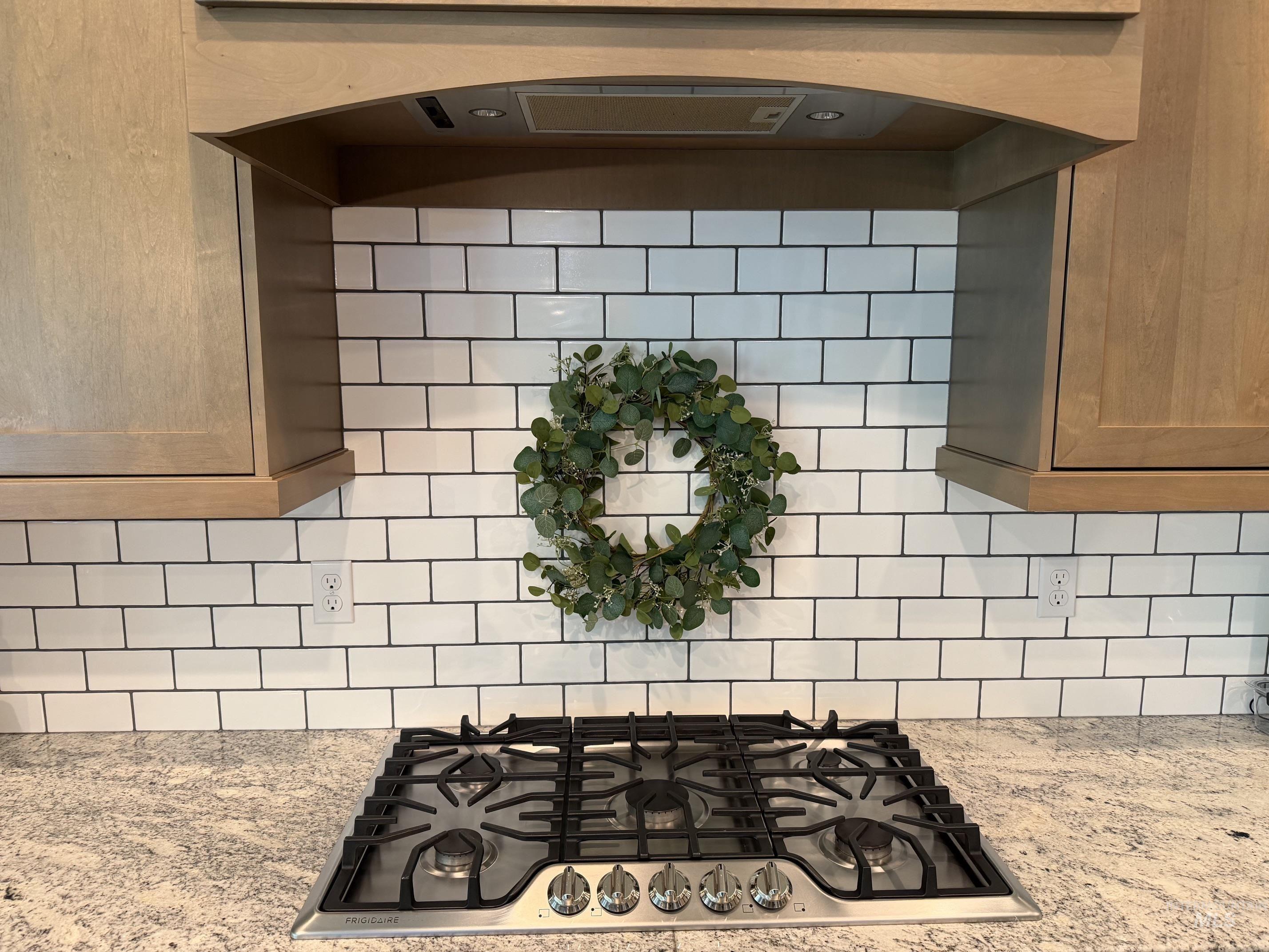 Kitchen featuring a gas cooktop, natural wood-finish cabinetry, a built-in range hood with integrated lighting, a white subway tile backsplash with dark grout, and speckled granite countertops