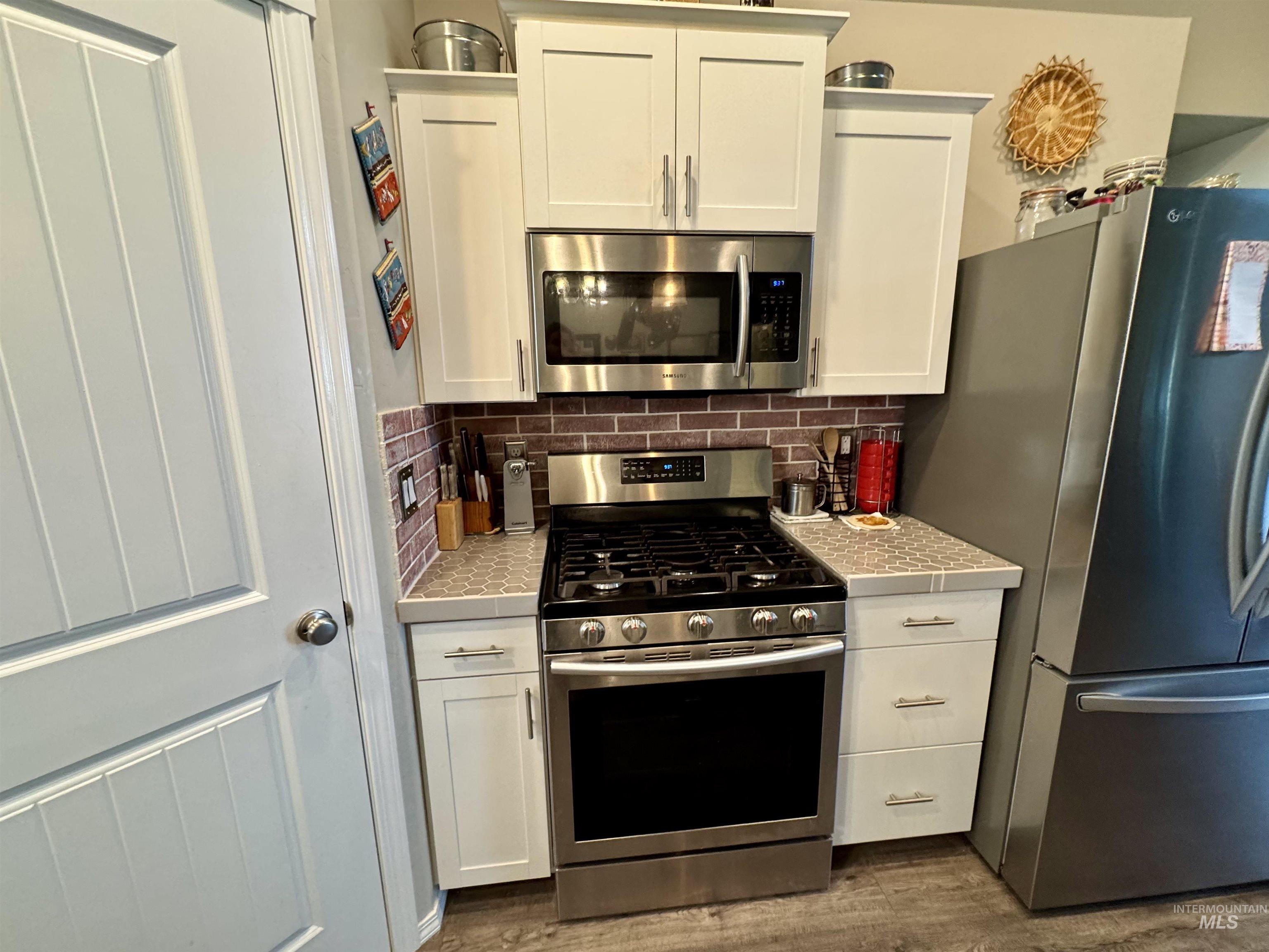 Kitchen with stainless steel appliances, white cabinetry, backsplash, tile countertops, and dark wood-type flooring