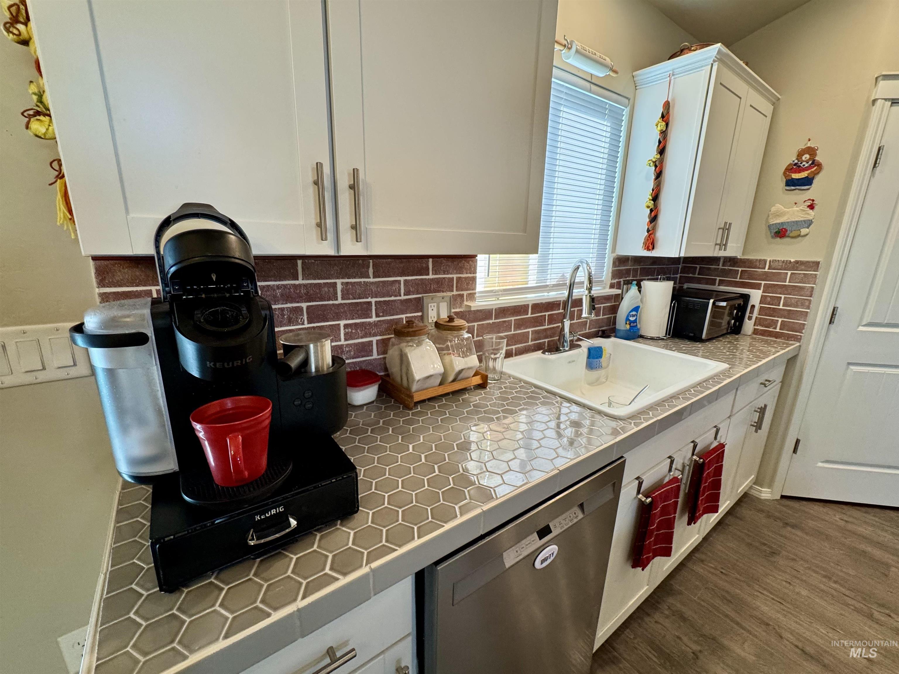 Kitchen with white cabinetry, stainless steel dishwasher, tile countertops, tasteful backsplash, and dark wood-type flooring