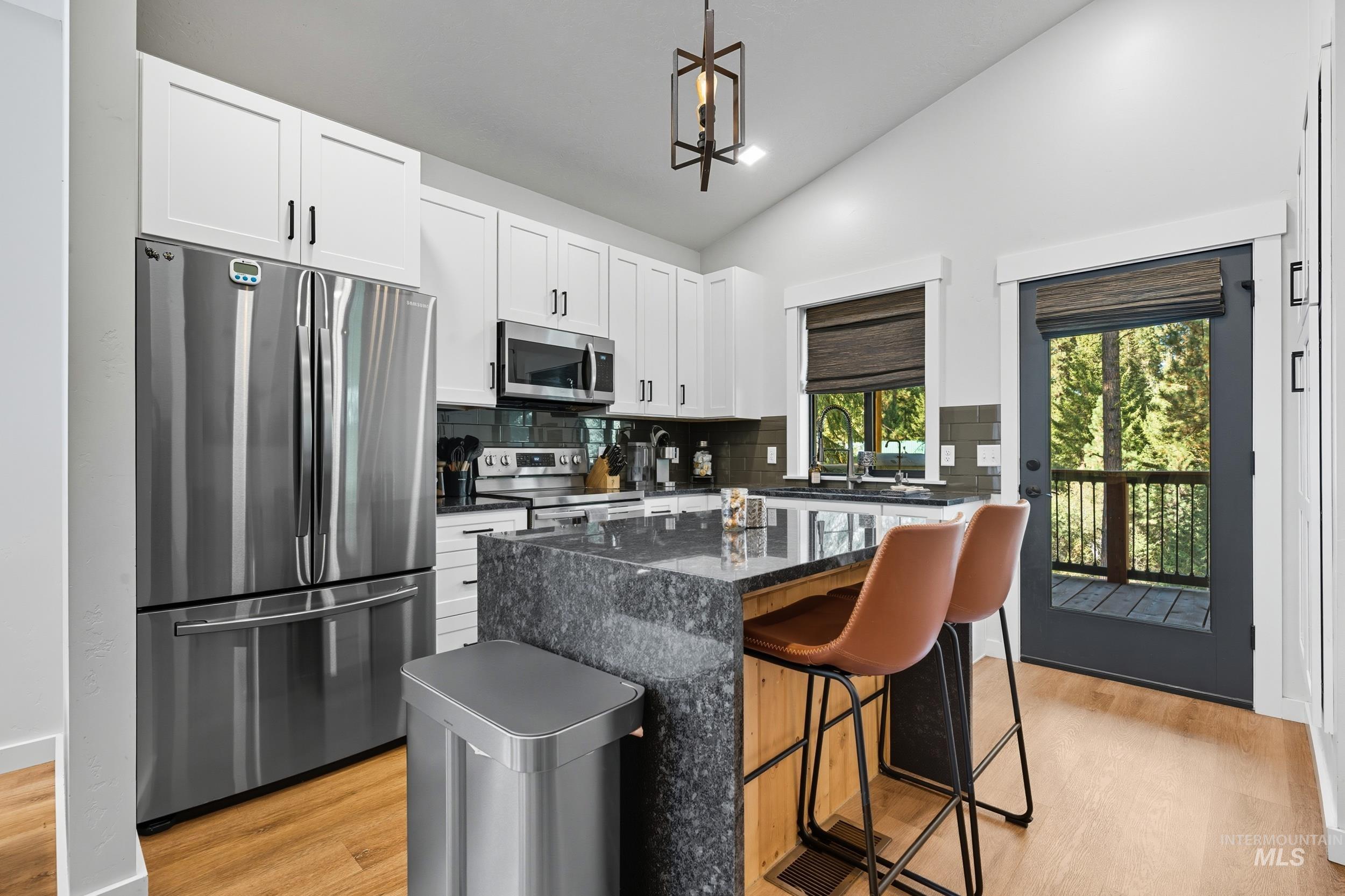 Kitchen with dark stone counters, appliances with stainless steel finishes, light wood-style floors, vaulted ceiling, and decorative light fixtures