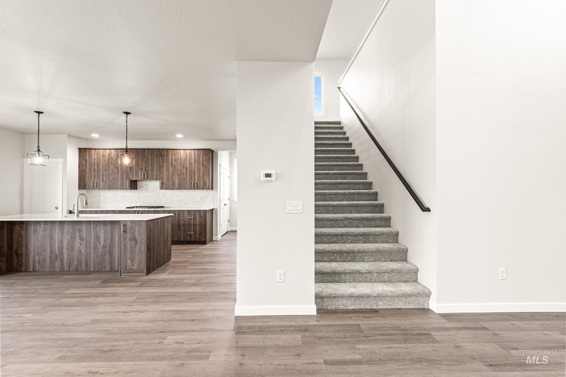 Open-concept living space with wood-finish flooring, a contemporary kitchen island with white countertop, dark wood-finish cabinetry, and a carpeted staircase with a black handrail