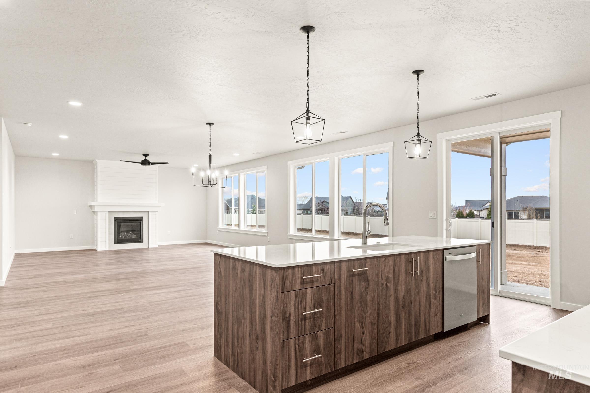 Open-concept living space featuring wood-finish flooring, a central island with a stainless steel sink, a built-in fireplace with a shiplap accent, multiple windows, and a sliding glass door to the exterior