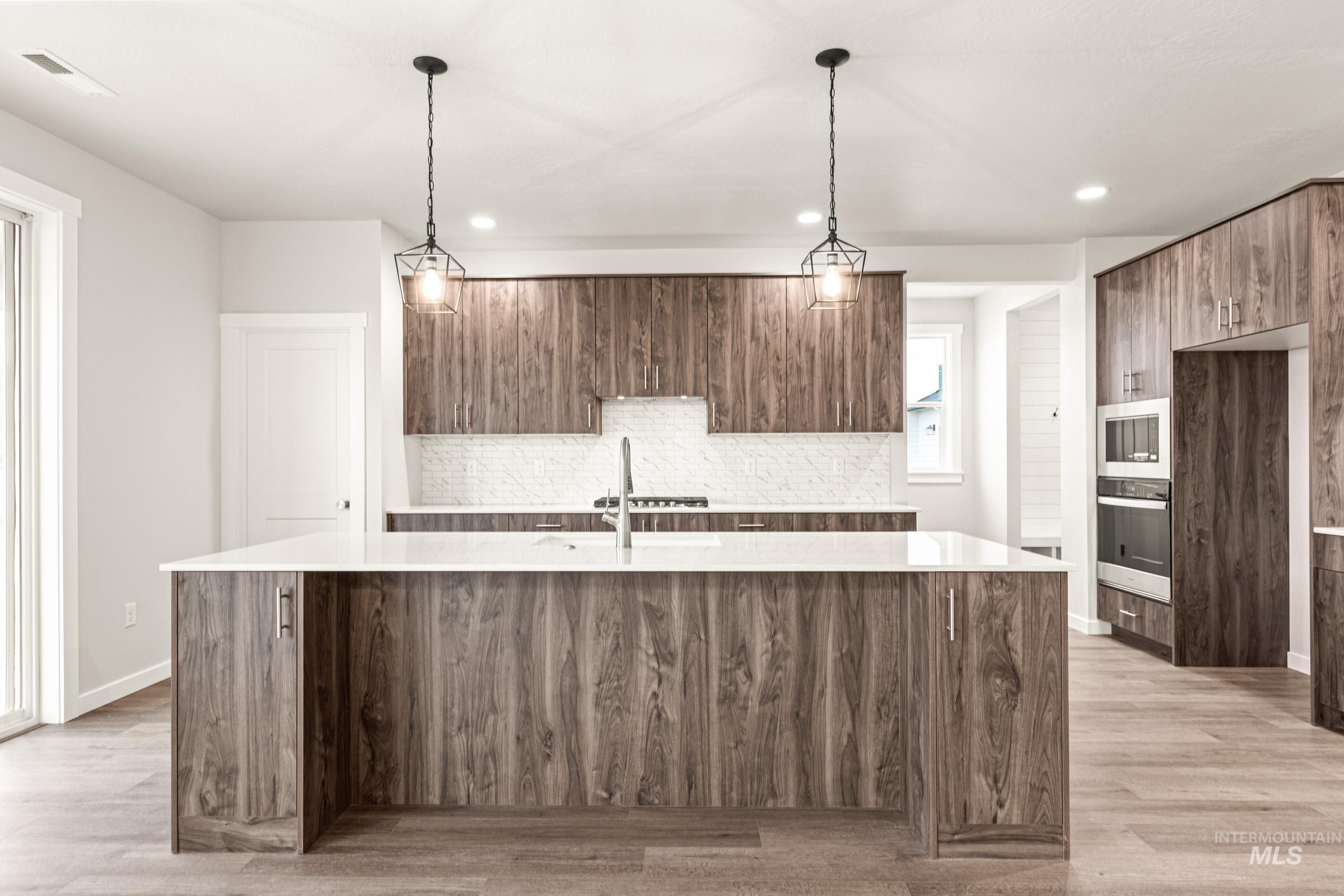 Spacious kitchen featuring a large central island with a white countertop, wood-finish cabinetry, built-in double ovens, and pendulum light fixtures