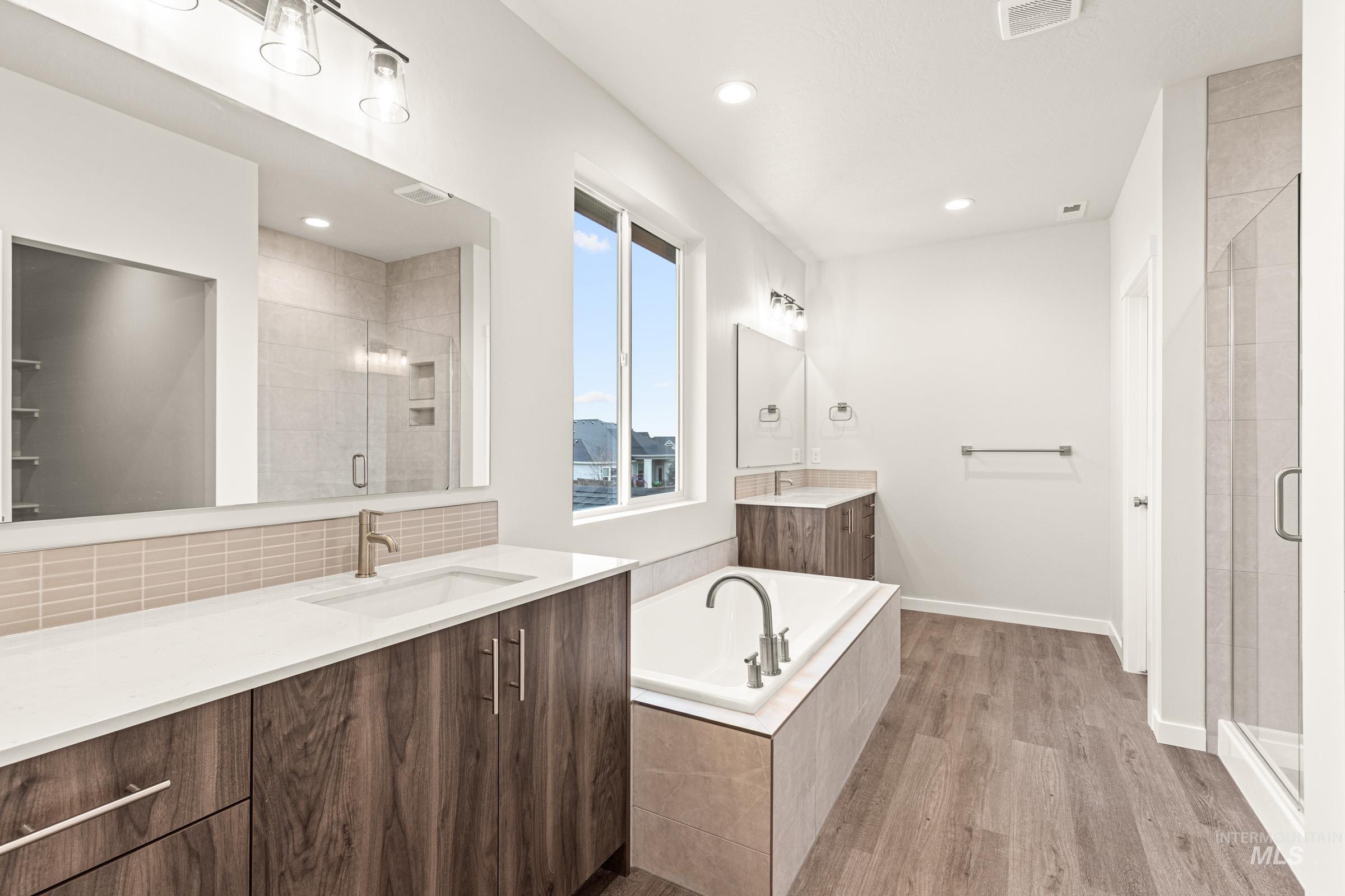Bathroom featuring a built-in soaking tub, a separate glass-enclosed shower, dual vanities with white countertops, wood-finish cabinetry, and large windows