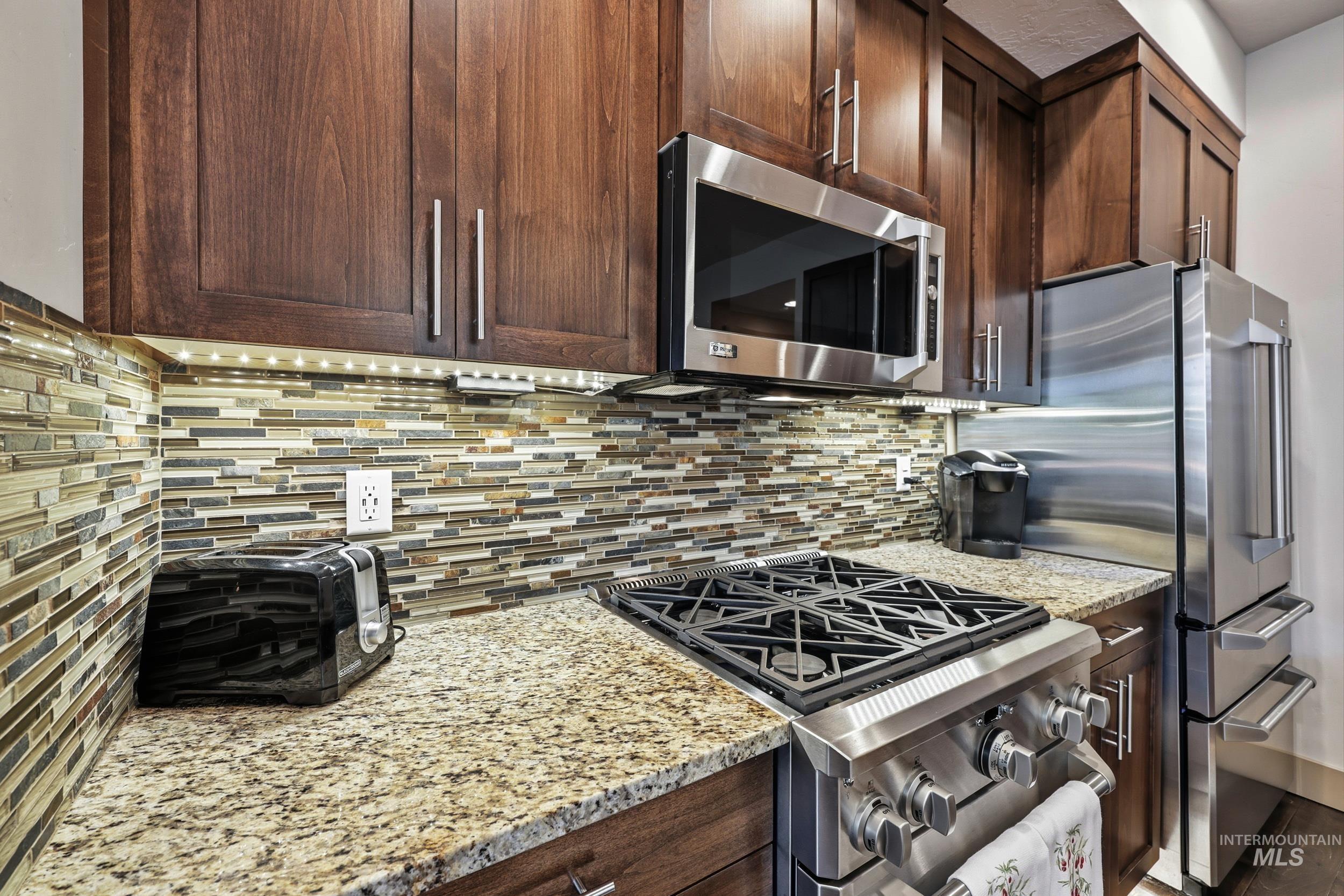Kitchen with appliances with stainless steel finishes, tasteful backsplash, light stone counters, and dark brown cabinets