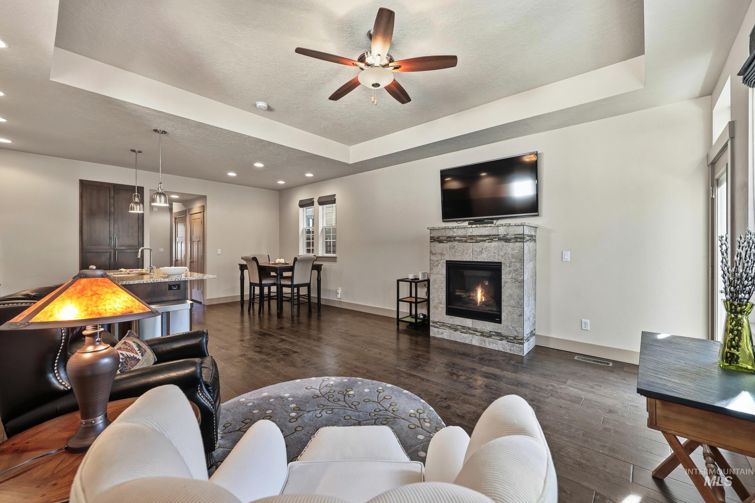 Living room featuring a tray ceiling, a glass covered fireplace, dark wood finished floors, ceiling fan, and recessed lighting