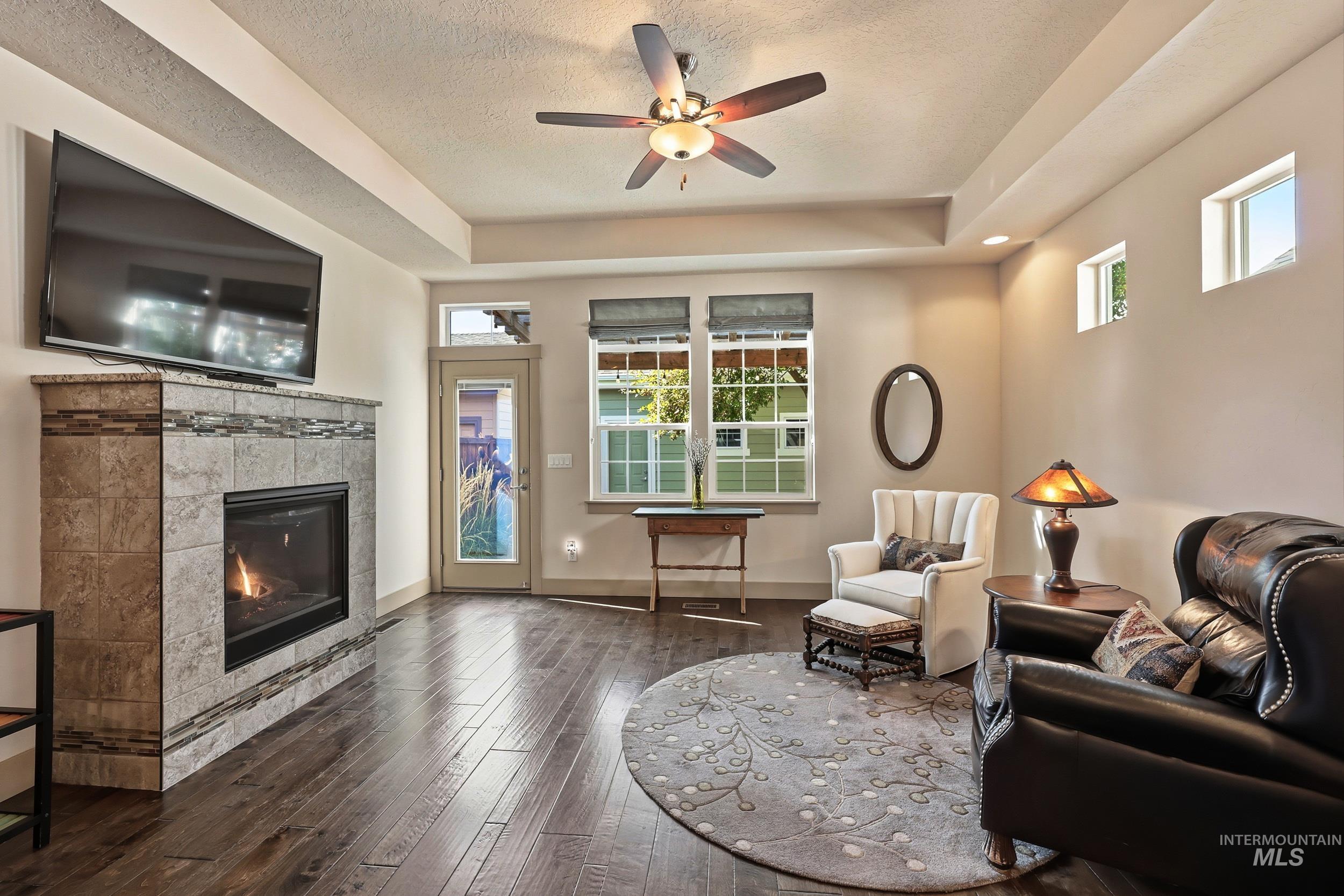 Living area with a raised ceiling, dark wood finished floors, a tiled fireplace, ceiling fan, and a textured ceiling