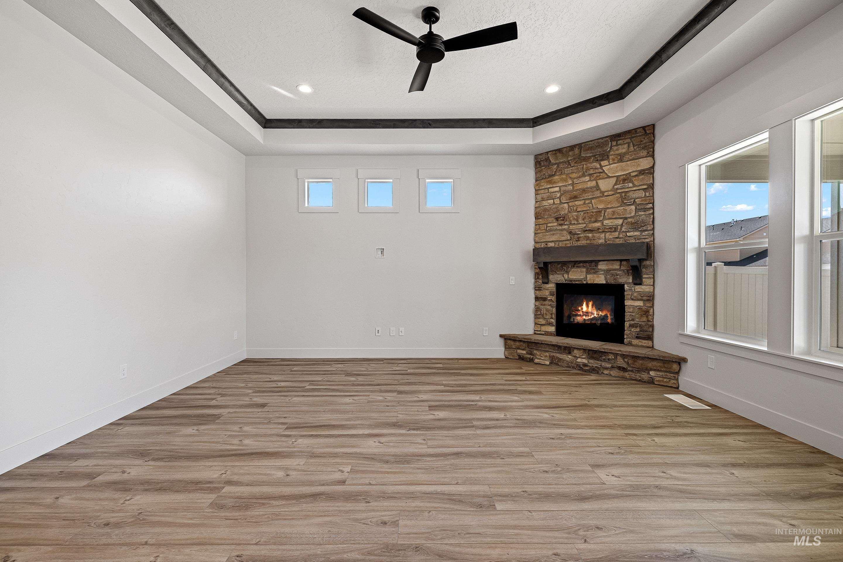 Unfurnished living room with ceiling fan, a raised ceiling, light wood finished floors, a stone fireplace, and recessed lighting