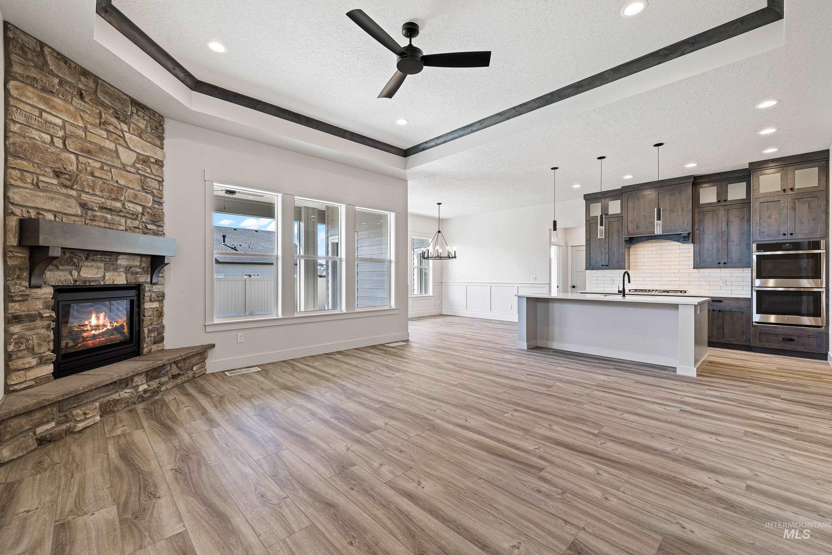 Unfurnished living room with a raised ceiling, light wood-type flooring, a chandelier, a stone fireplace, and ceiling fan